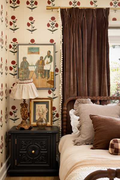 close up photo of a corner of a bedroom with cream and red floral wallpaper on the walls, a painting of men hung on the wall and brown velvet curtains hung in front of an illuminated window, a brown wooden frame bed in front of the window with brown pillows and white bedding, a black night stand with a gold lamp sitting on it next to the bed