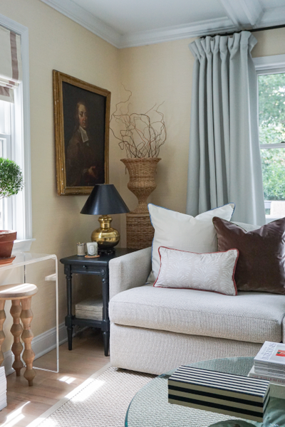 close up picture of the corner of a living room with cream colored walls, a portrait of a man in a gold frame hung on the wall, pale blue grey curtains hung in front of a window with a white couch in front of the window with white, brown velvet, and neutral botanical stripe pillows on the couch, a navy blue side table next to the couch with a gold and navy lamp on the table