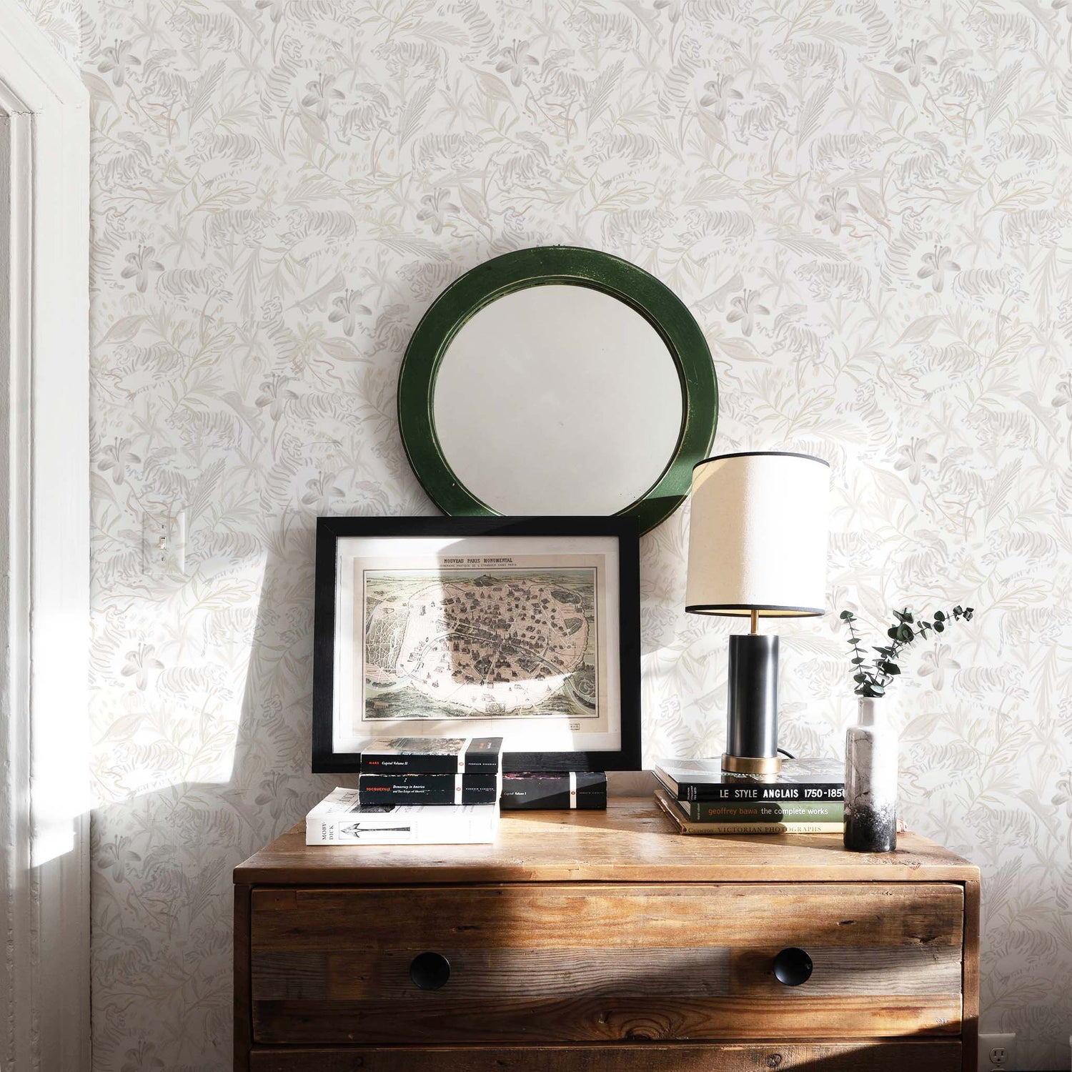 Corner styled with Beige Chinoiserie Tiger Printed Wallpaper by wooden dresser with black and white lamp on top of stacked books next to picture frame below circular green mirror