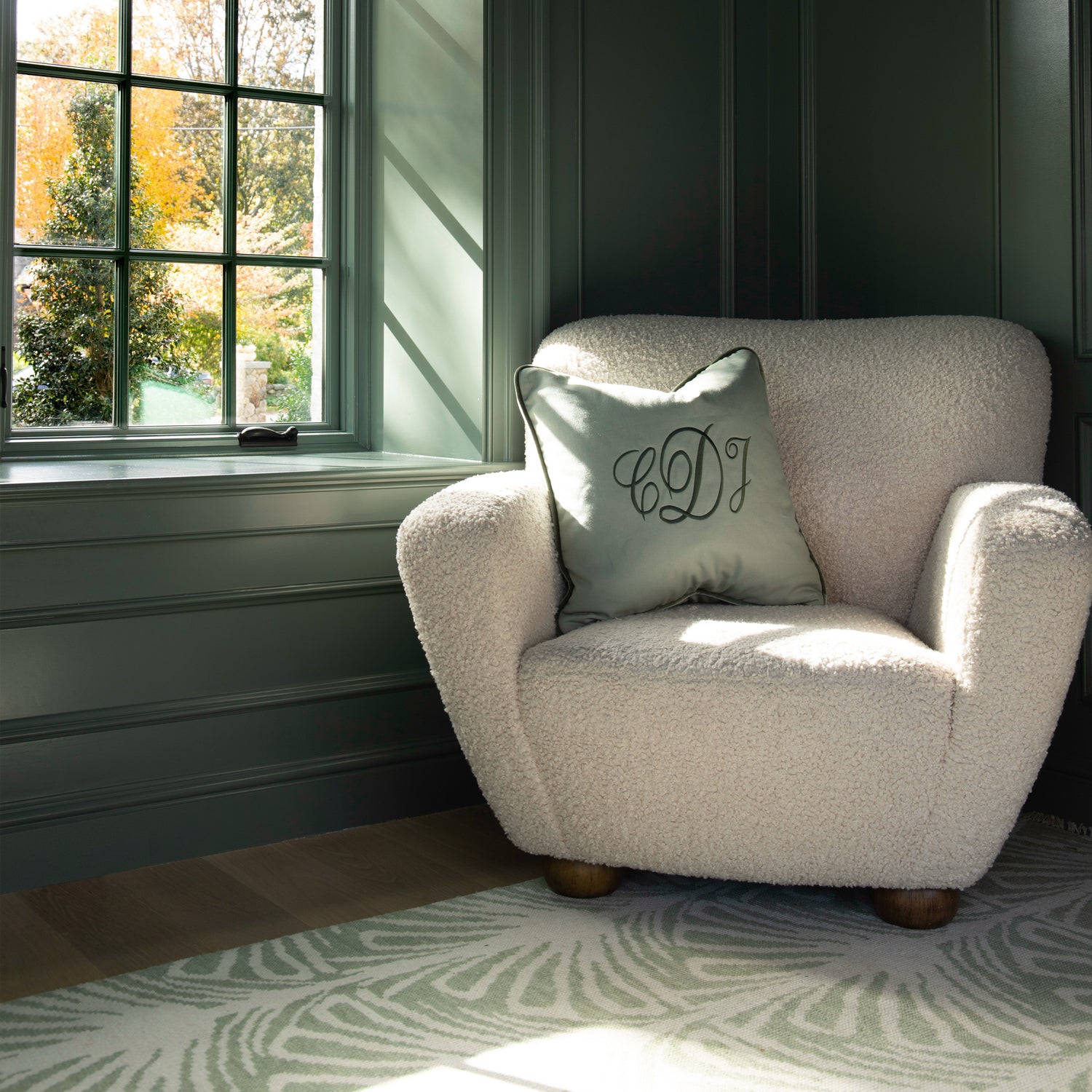 Corner close-up of white sofa chair with Sage Green Monogrammed Pillow on top next to illuminated window and Sage Green Printed Rug on wooden floor