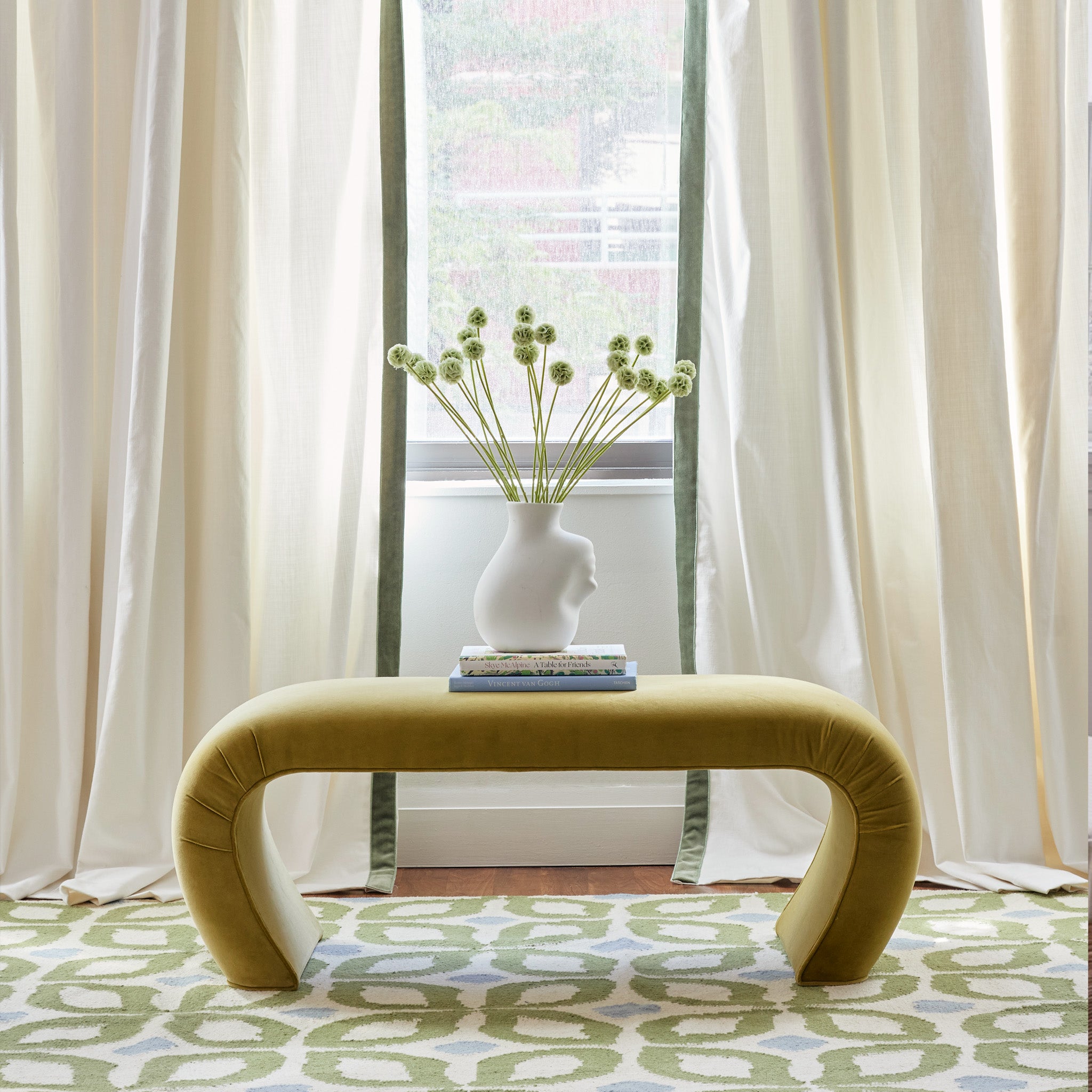 white curtains with a sage green velvet band hung in front of an illuminated window with a geometric green and blue rug and a dark yellow table with books and a vase on it 