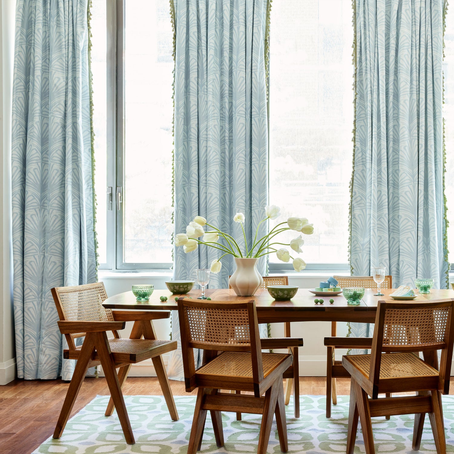 Wooden dining room table styled with white tulips in white vase in the center and Moss Green Geometric Printed Rug below in front of Sky Blue Palm Printed Curtains