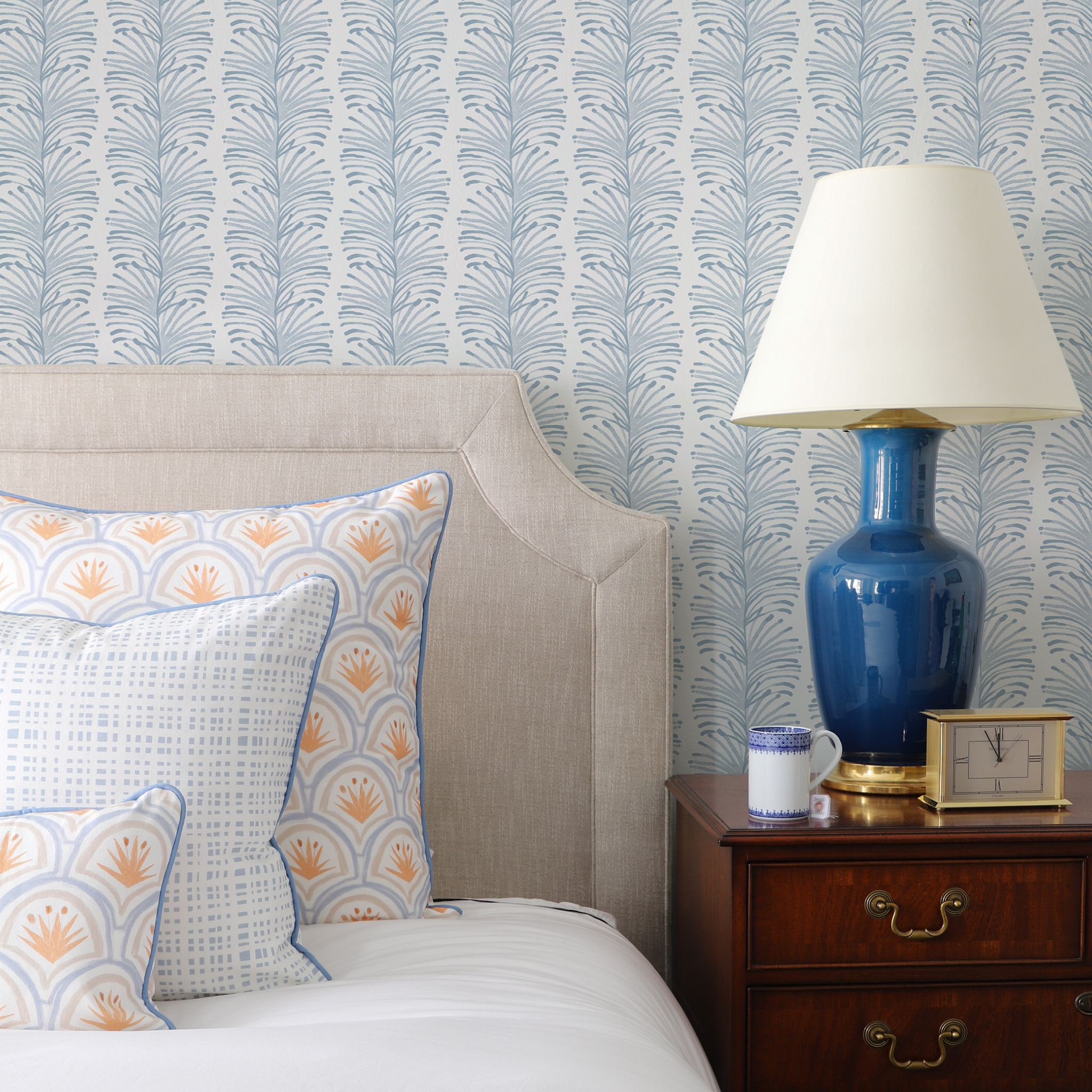 Bedroom corner with beige upholstered headboard, patterned pillows, blue ceramic lamp, wooden nightstand, and leaf-print wallpaper.