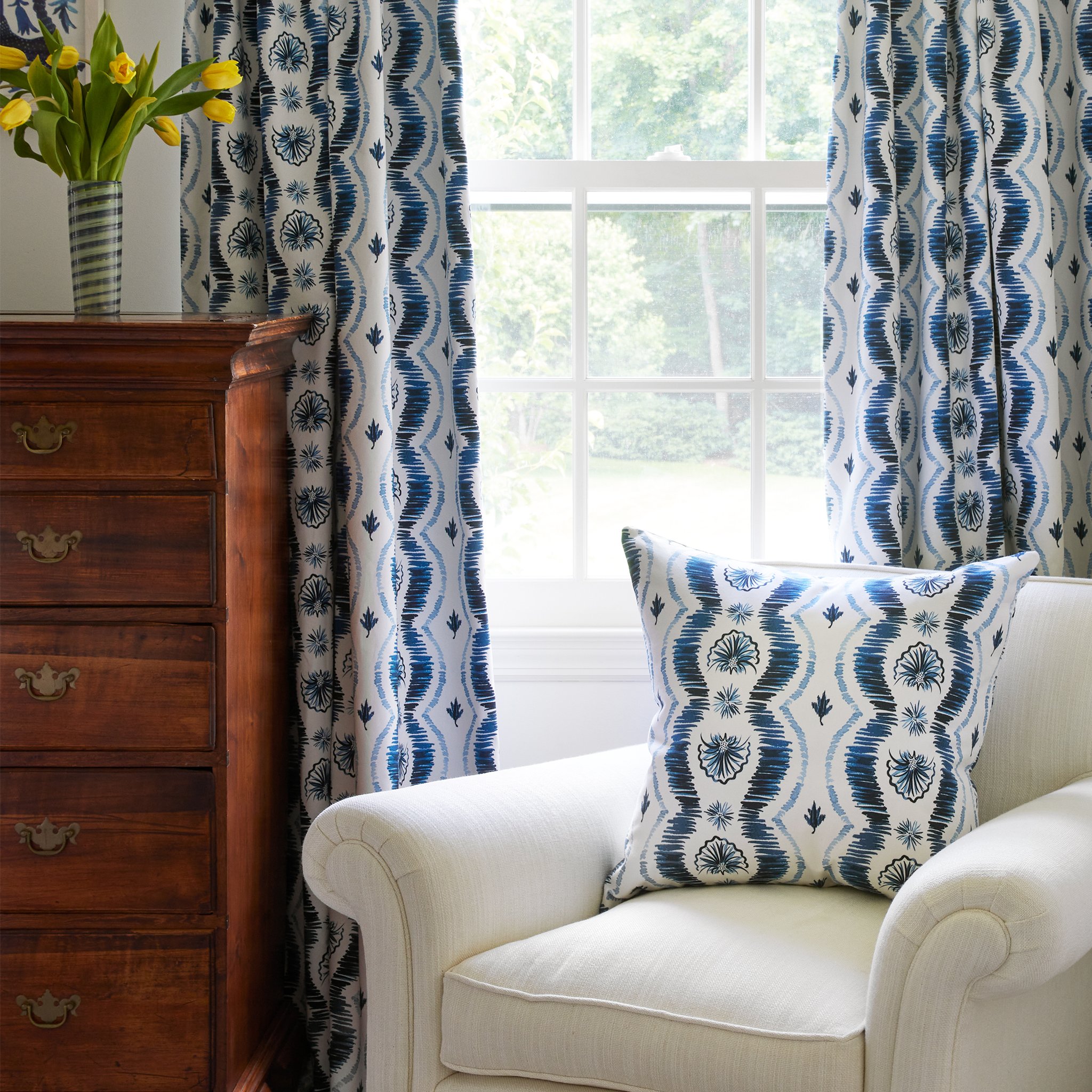 Blue Ikat pillow on couch with Blue Ikat striped curtains by a nightstand with a flower vase