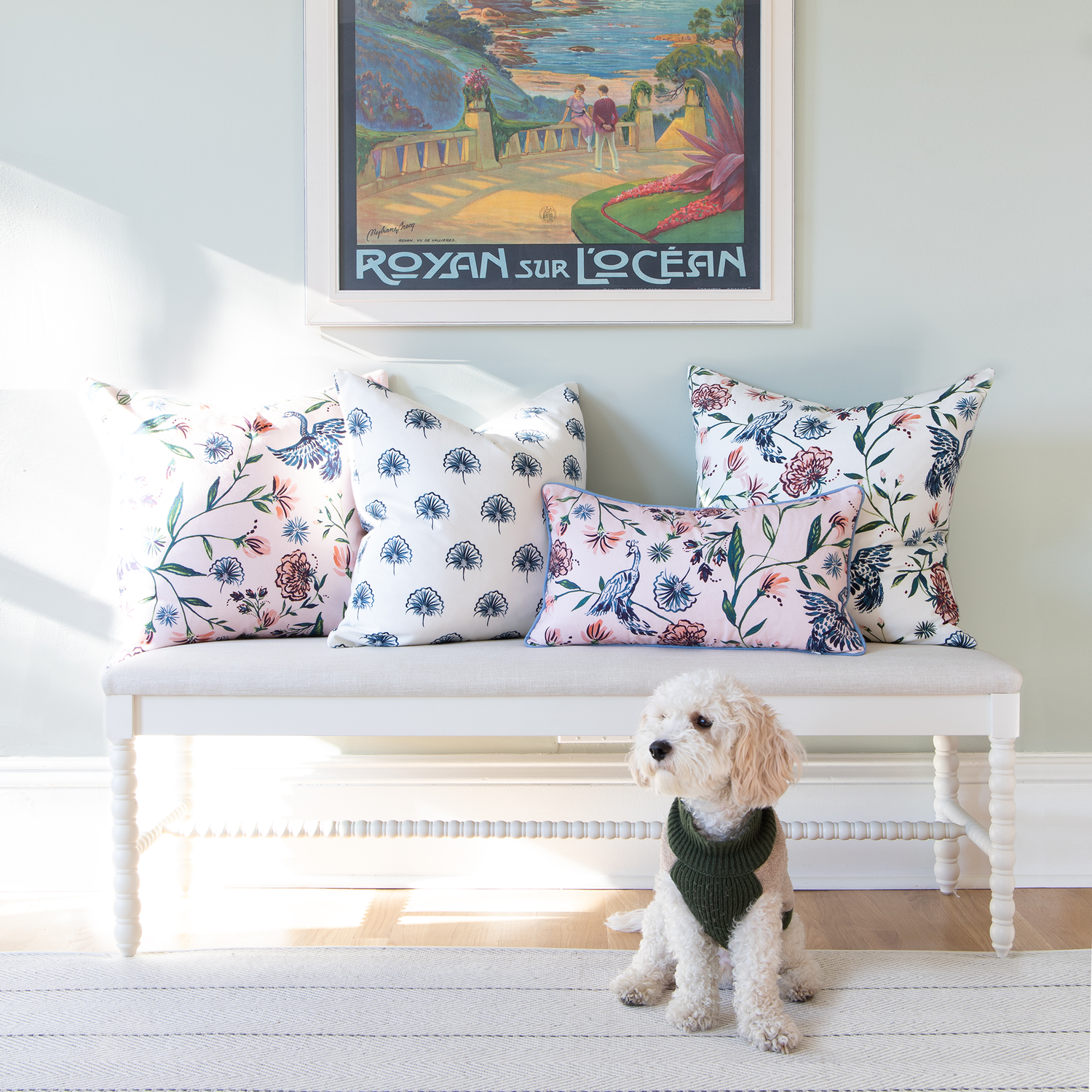 Space styled with Cream Chinoiserie, Rose Chinoiserie, and Floral Navy pillows on white bench with dog wearing a green vest in front of wall with painting