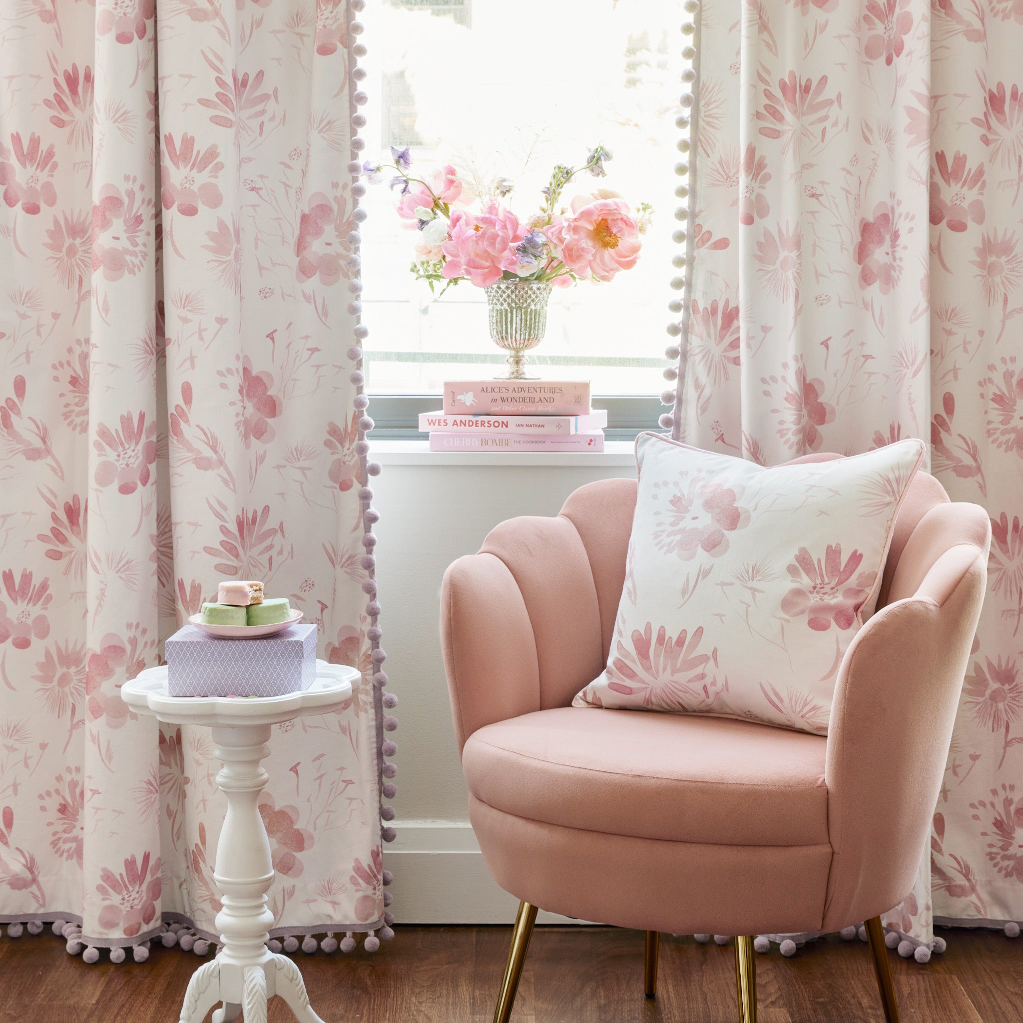 Pink Sofa Chair with Pink Floral Pillow on top and burgundy velvet loafers on the ground by a white small table with a box and dessert plate on top in front of Pink Floral Curtains with books and flower vase on top