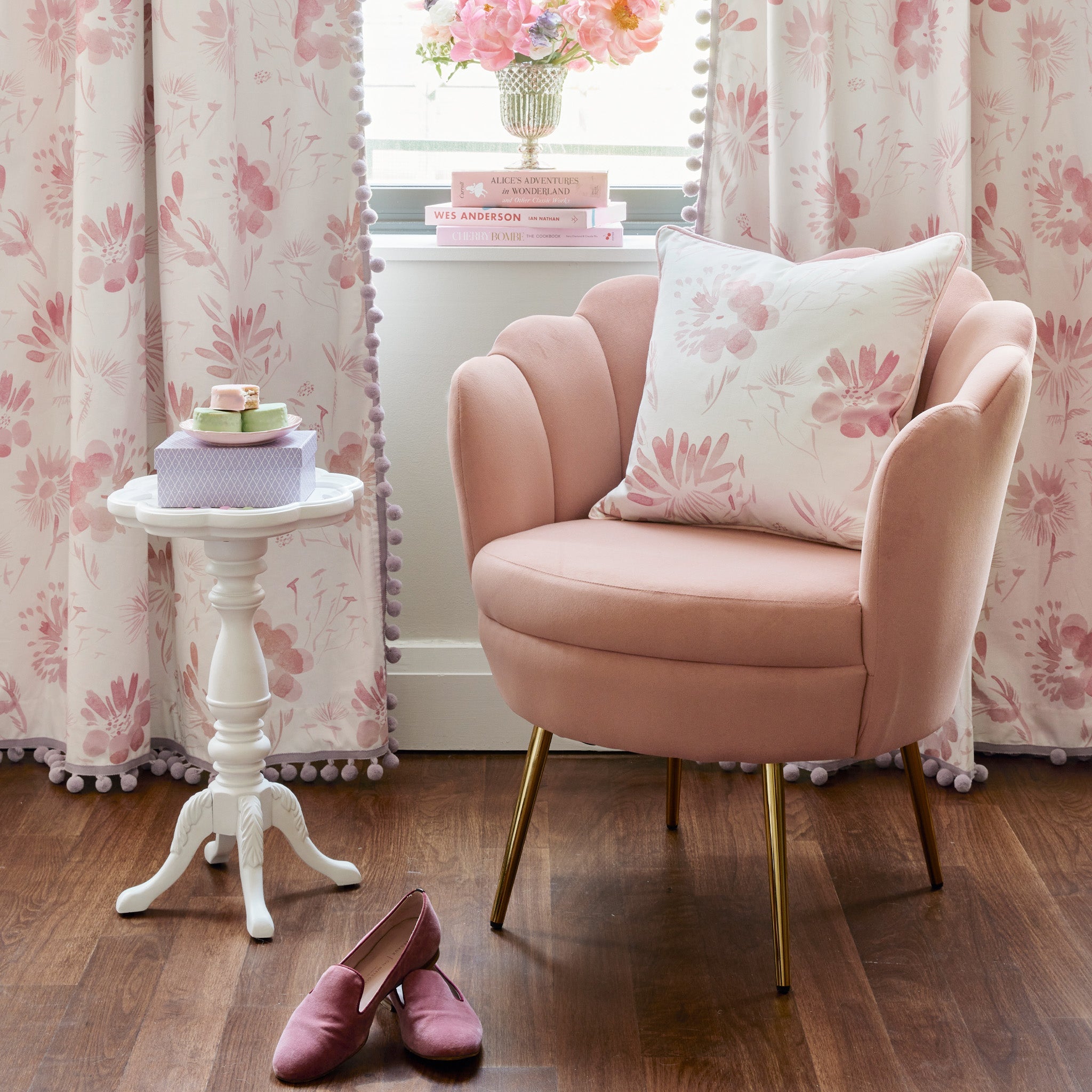 Pink Sofa Chair with Pink Floral Pillow on top and burgundy velvet loafers on the ground by a white small table with a box and dessert plate on top in front of Pink Floral Curtains with books and flower vase on top