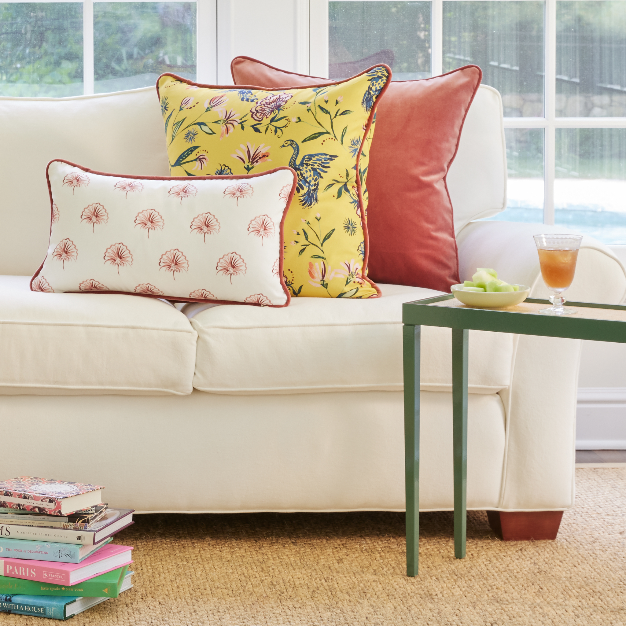 White Couch with Floral Pink, Yellow Chinoiserie, and Coral Velvet pillows with Green table with plate on top with two stacks of books on the floor