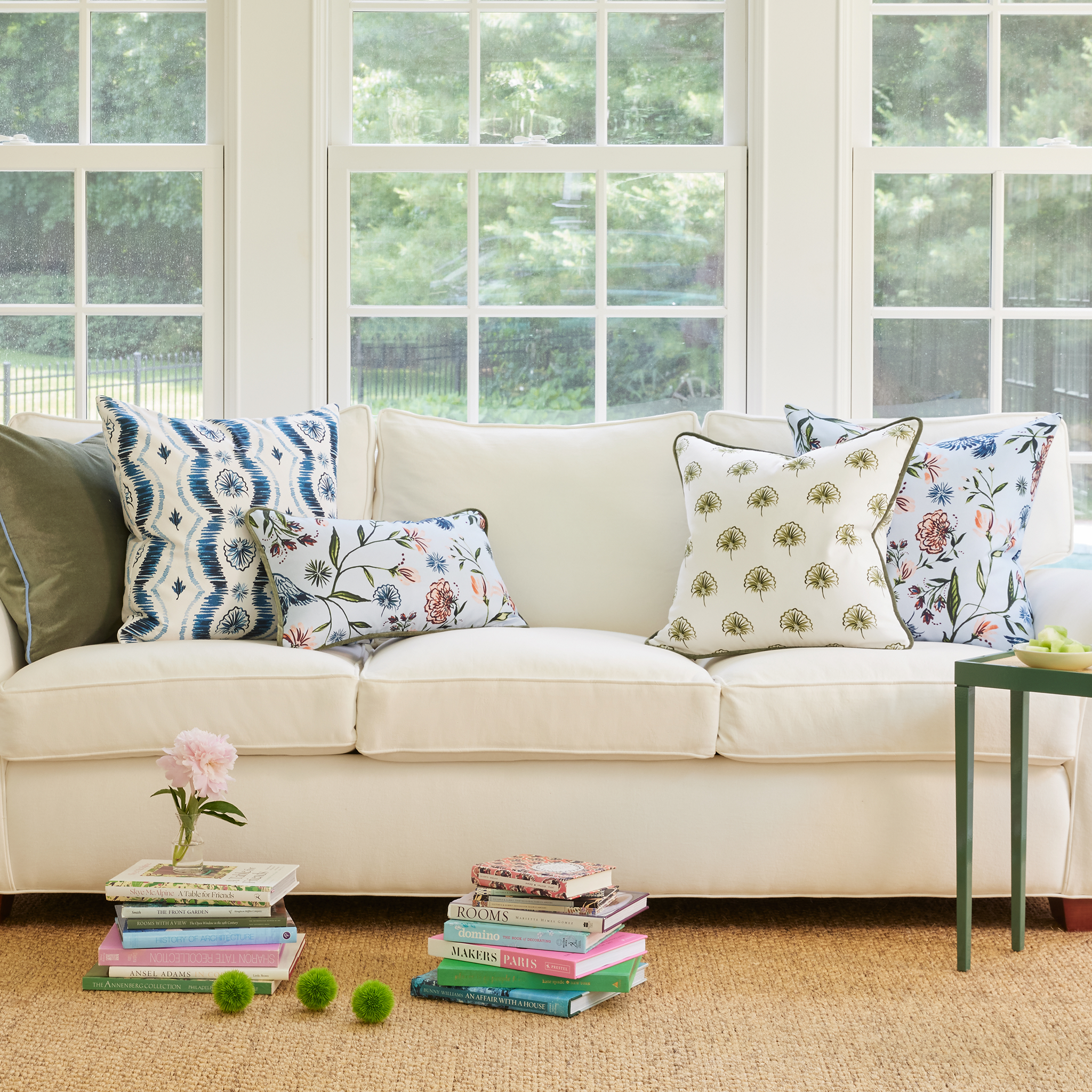 White couch on living room with fern green pillow, blue ikat pillow, daphne powder pillow, and green floral pillow by two stacks of books on the floor with one stack having a pink flower on top with white framed window in the back