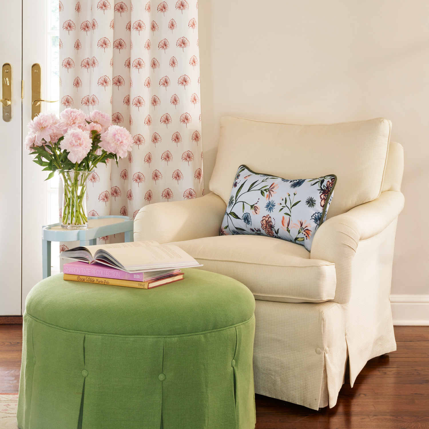 Living room styled with Blue Chinoiserie pillow on beige sofa chair with Rose Floral Curtain hanging next to it and pink flowers in transparent vase on top of small blue table in the back of green circular couch with books stacked on top