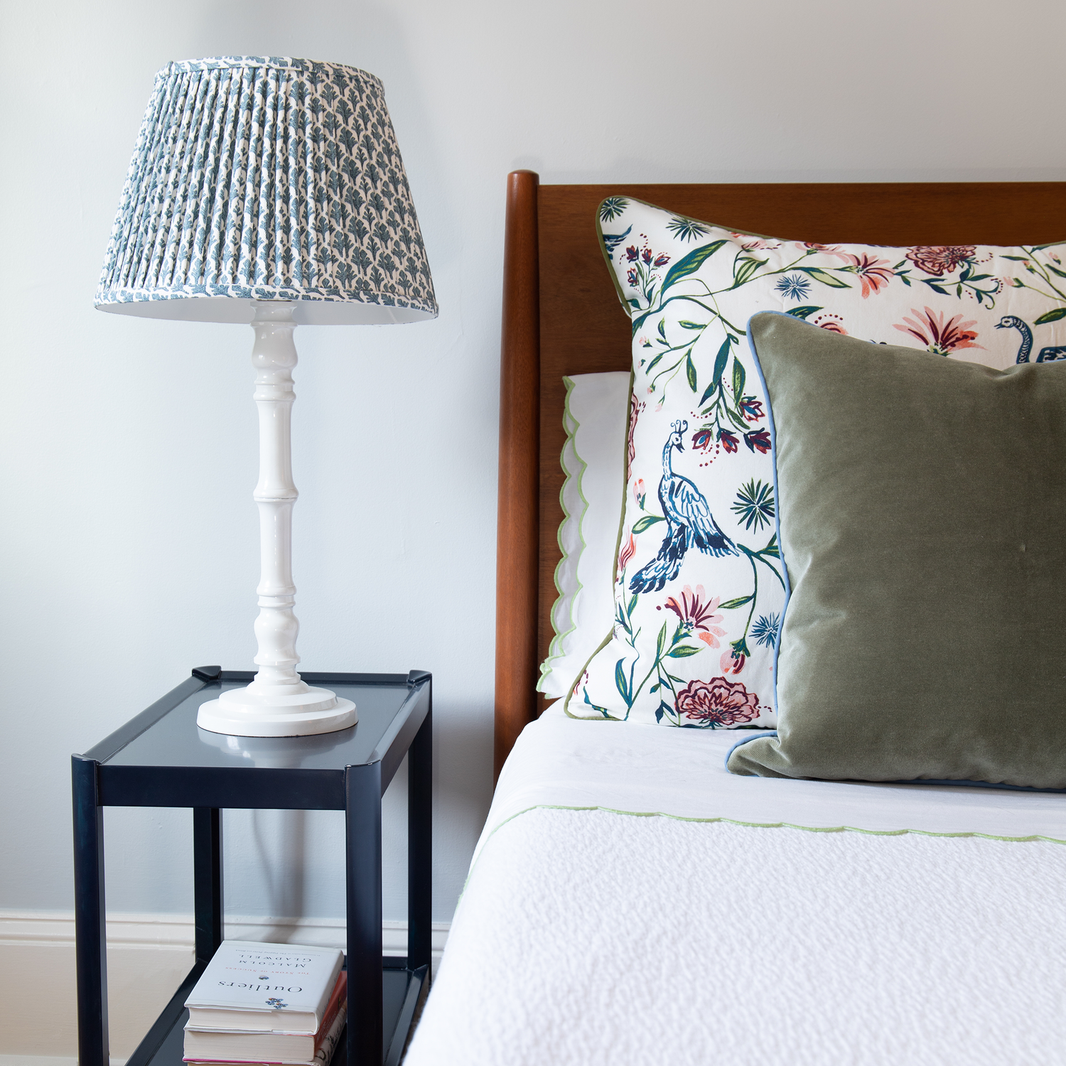 Bedroom styled with white bed with wooden headboard and Cream Chinoiserie and Fern Green Pillows next to navy night stand with white lamp and books stacked underneath