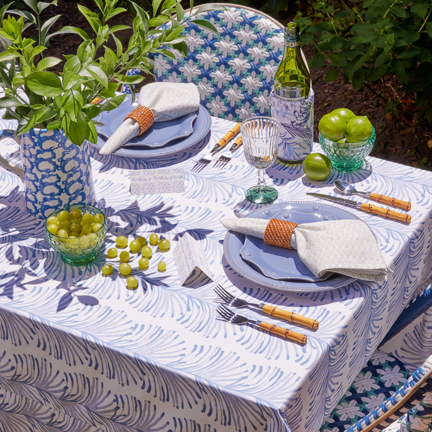 Table styled with Sky Blue Botanical Stripe Tablecloth and Moss Green Geometric Napkins on blue plates and silverware next to them with fruit cups to the side and plants in blue and white vase