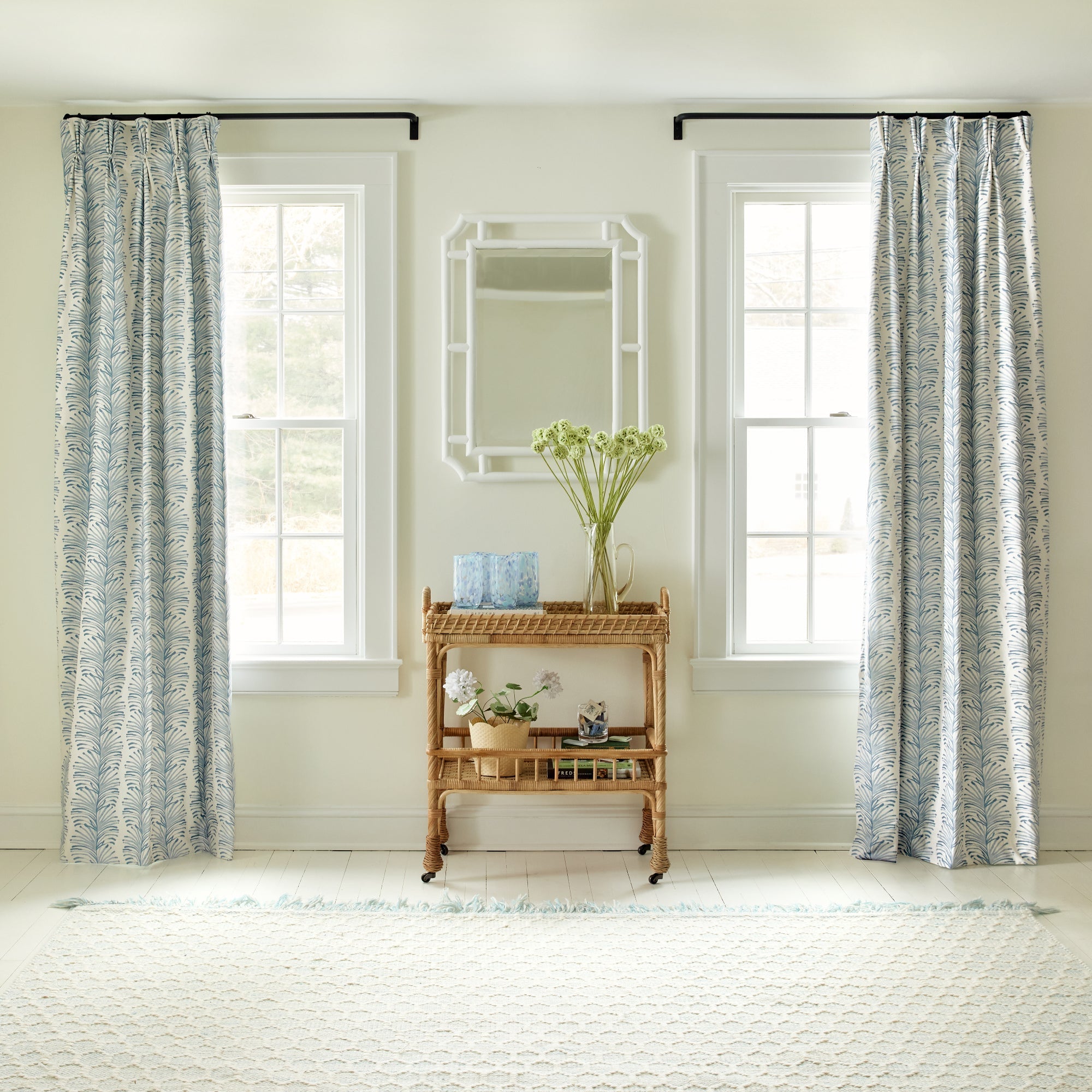 Windows styled with Sky Blue Botanical Stripe Printed Curtains and brown cart in the middle with green flowers in vase on top and white mirror on wall in between windows