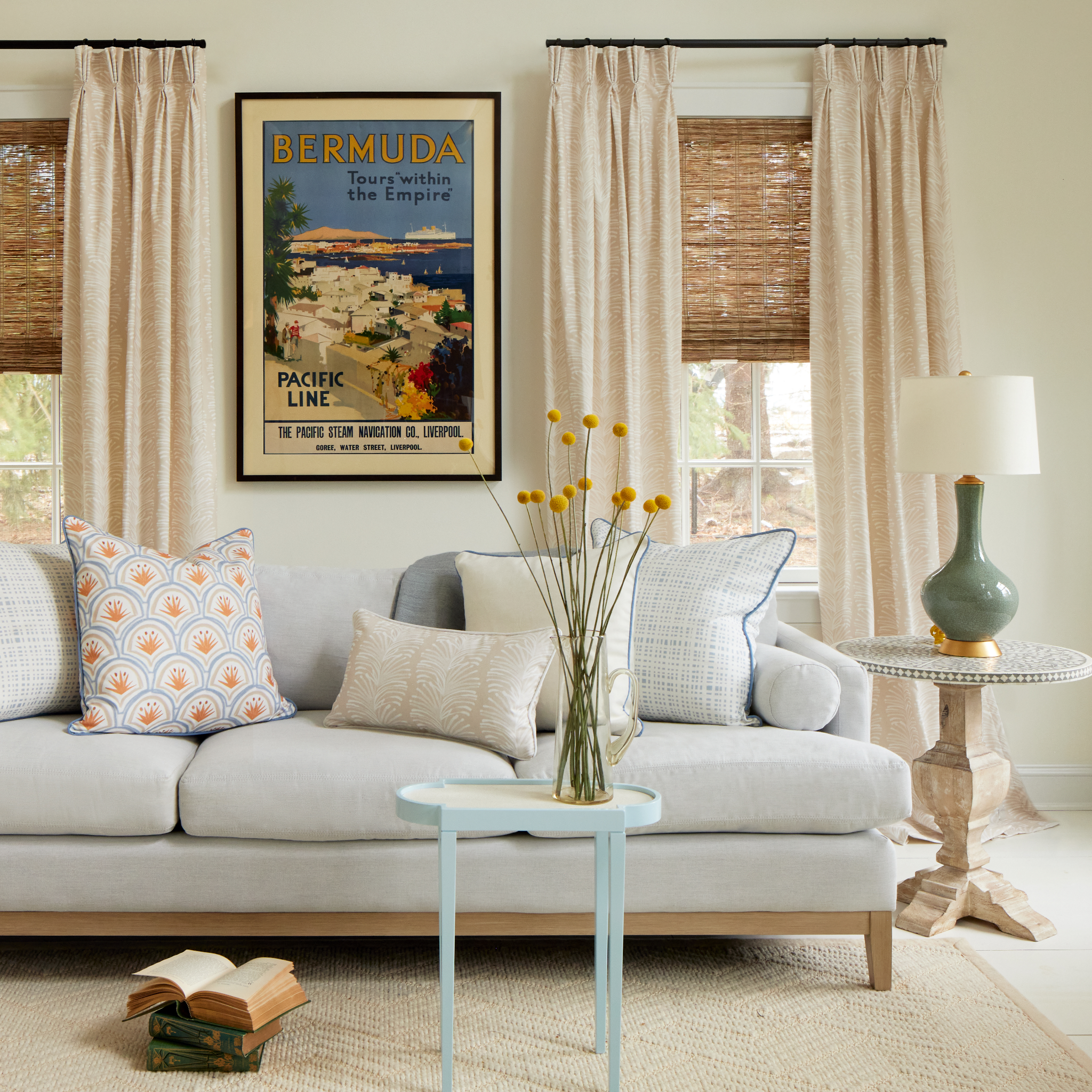 Living room styled with two sky blue gingham printed pillows, Art Deco Palm Pattern Printed Pillow, Beige Botanical Stripe Printed Lumbar, and Light brown Pillow on top of grey couch with Beige Botanical Stripe set of two curtains, books stacked on floor and blue and white circular table with yellow flowers in clear vase