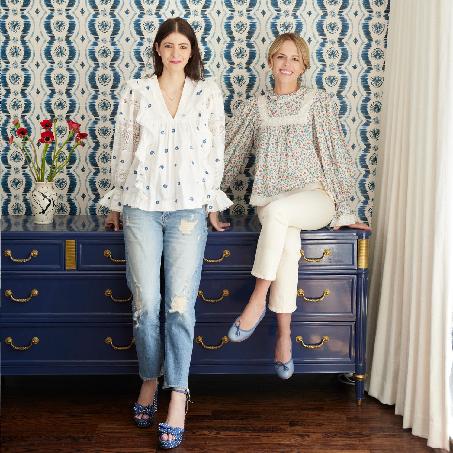 Pepper Home's Founders, Kelsey and Erin, on top of a blue dresser with flower vase in front of Blue Ikat Striped Pattern Wallpaper and white curtains on the far left