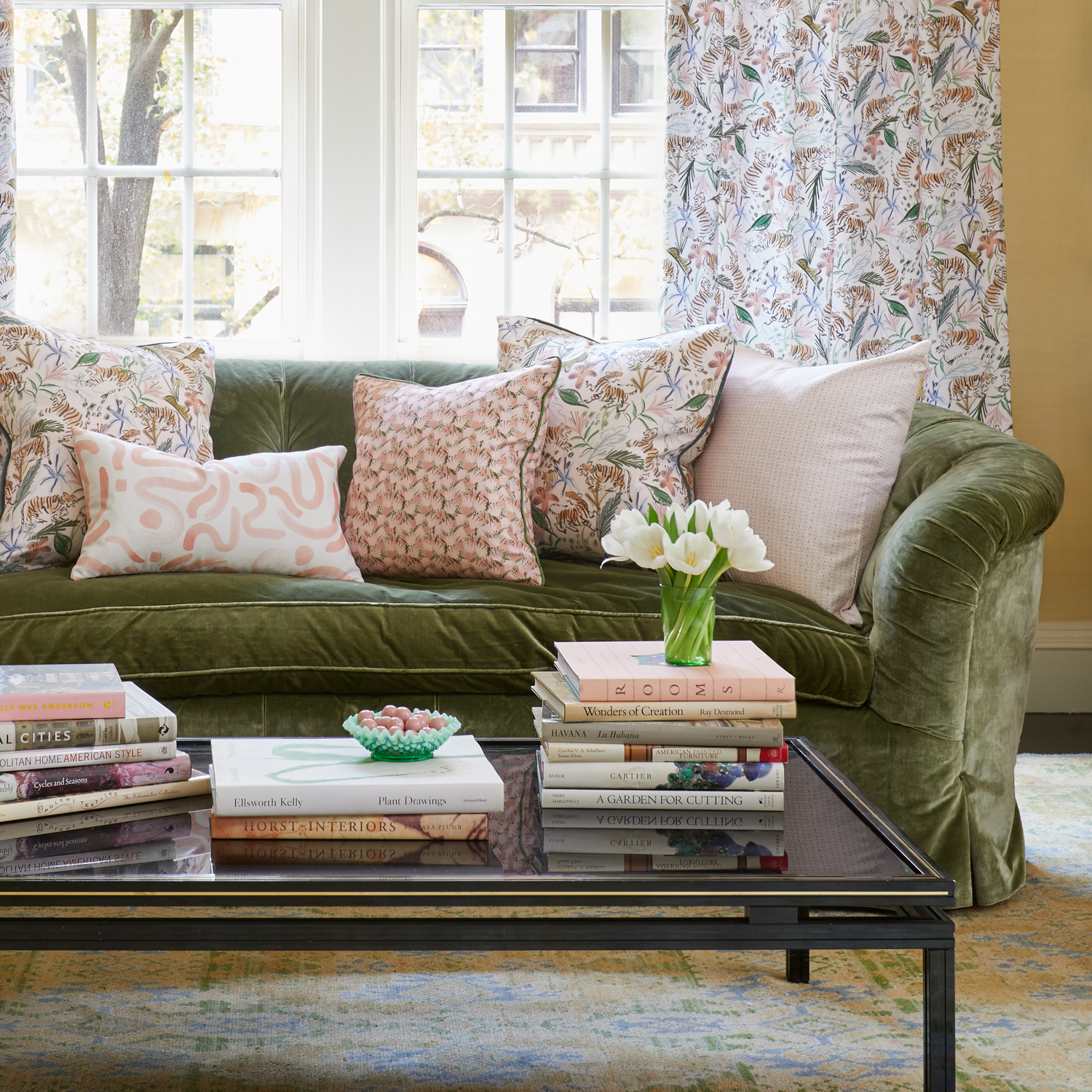 Living room styled with a Pink Geometric printed pillow, Pink Chinoiserie printed pillow, Pink Floral printed pillow, and Pink Graphic printed lumbar on Fern Green Velvet couch by coffee table with stacked books and white tulips in vase in front of an illuminated window with Cream Chinoiserie printed curtains 