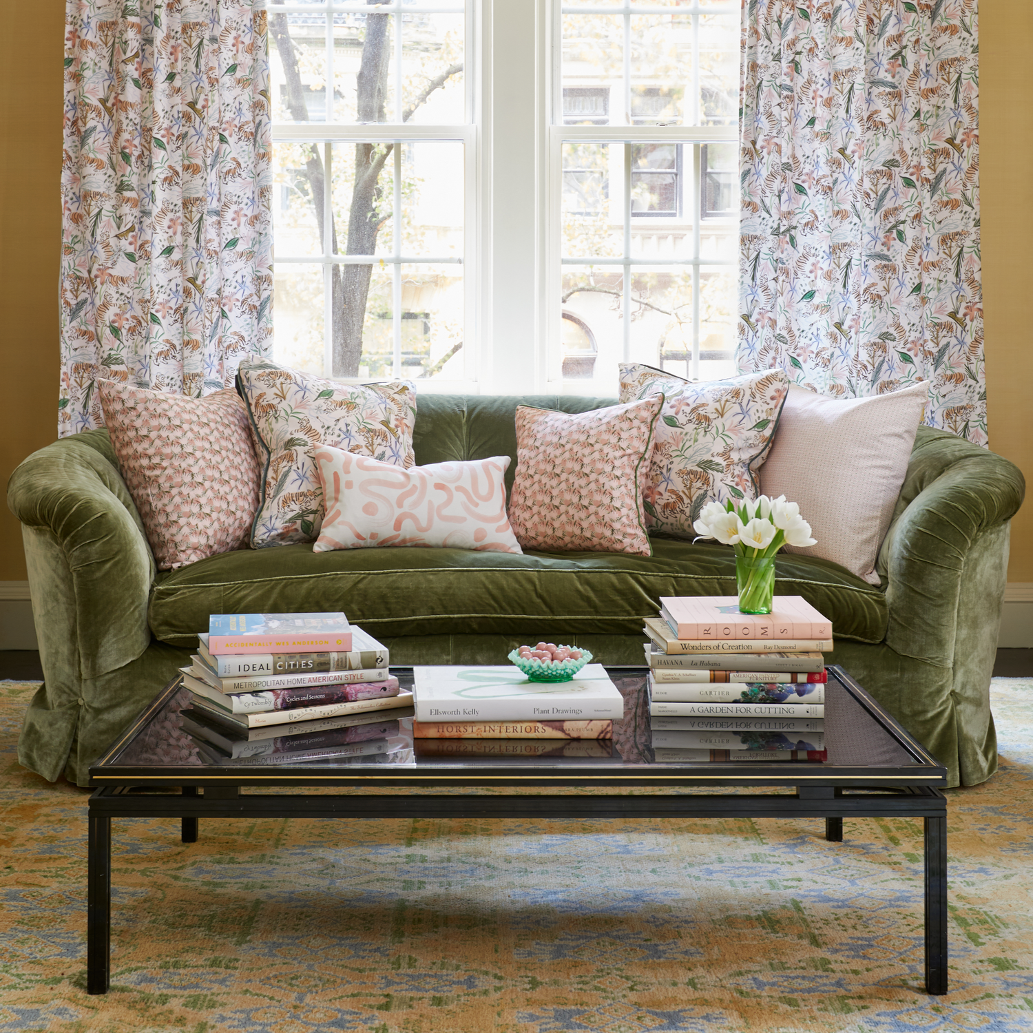 Living room styled with a Pink Geometric printed pillow, Pink Chinoiserie printed pillow, Pink Floral printed pillow, and Pink Graphic printed lumbar on Fern Green Velvet couch by coffee table with stacked books and white tulips in vase in front of an illuminated window with Pink Chinoiserie Tiger printed curtains