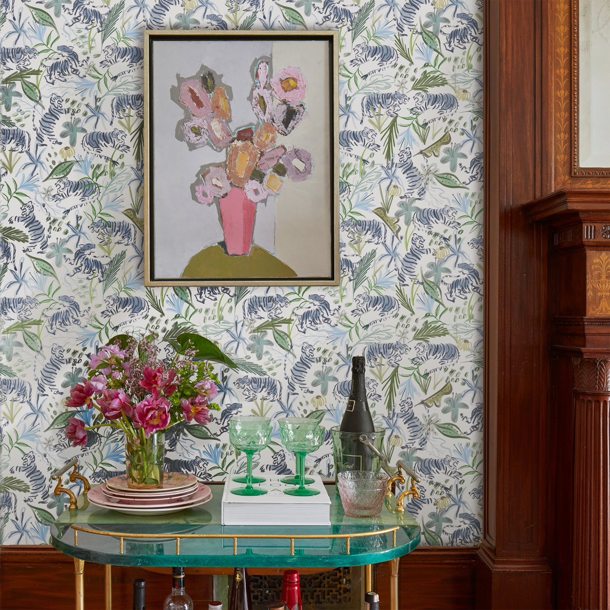 Living room space styled with Green Tiger Printed Wallpaper with artwork hung on top of green bar cart decorated with pink flowers in transparent vase on top of pink stacked plates next to green glasses