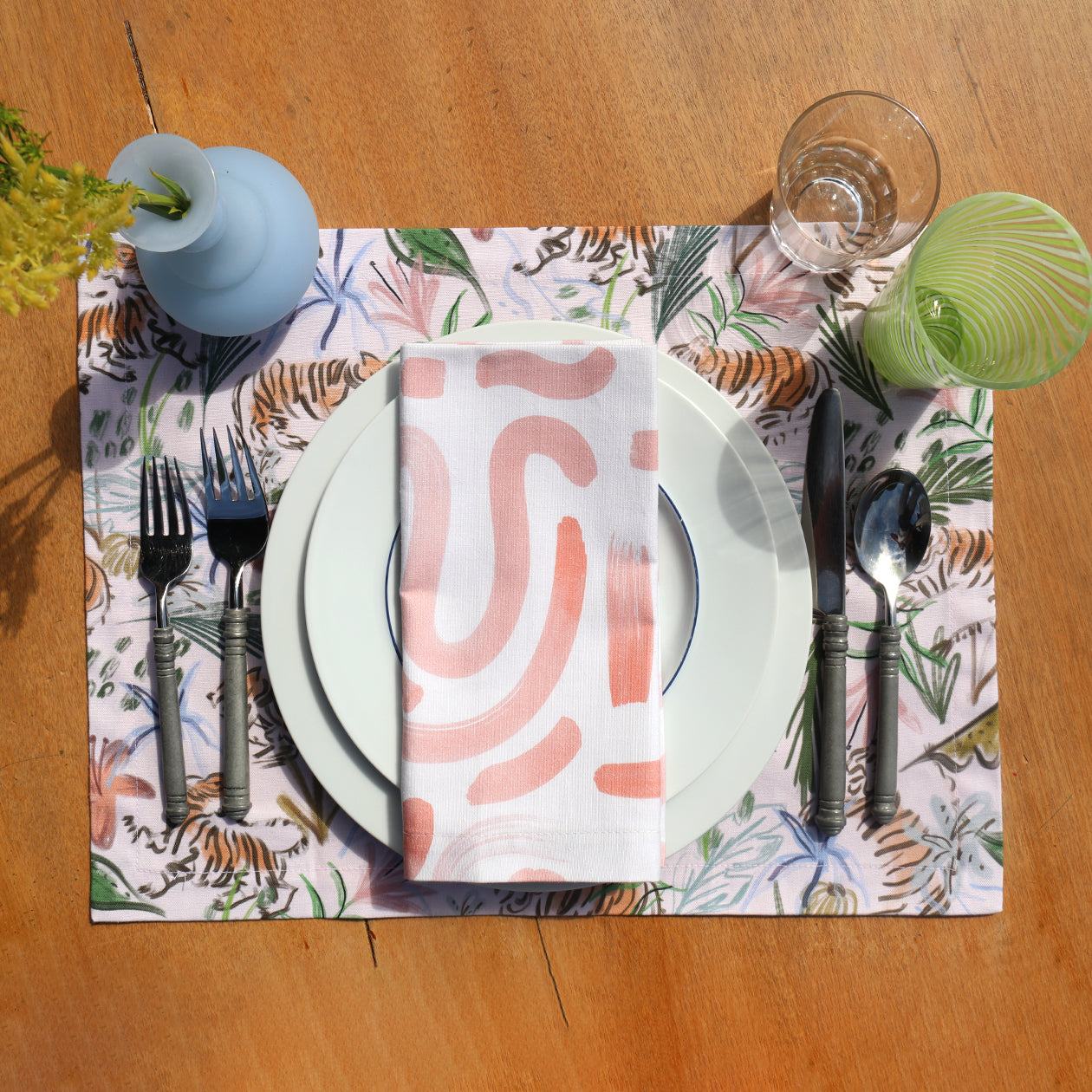 Close-up of table setting styled with Pink Chinoiserie Tiger Printed Placemat under two white plates with Pink Graphic Printed Napkin on top by silverware and two glass cups next to blue vase with flowers