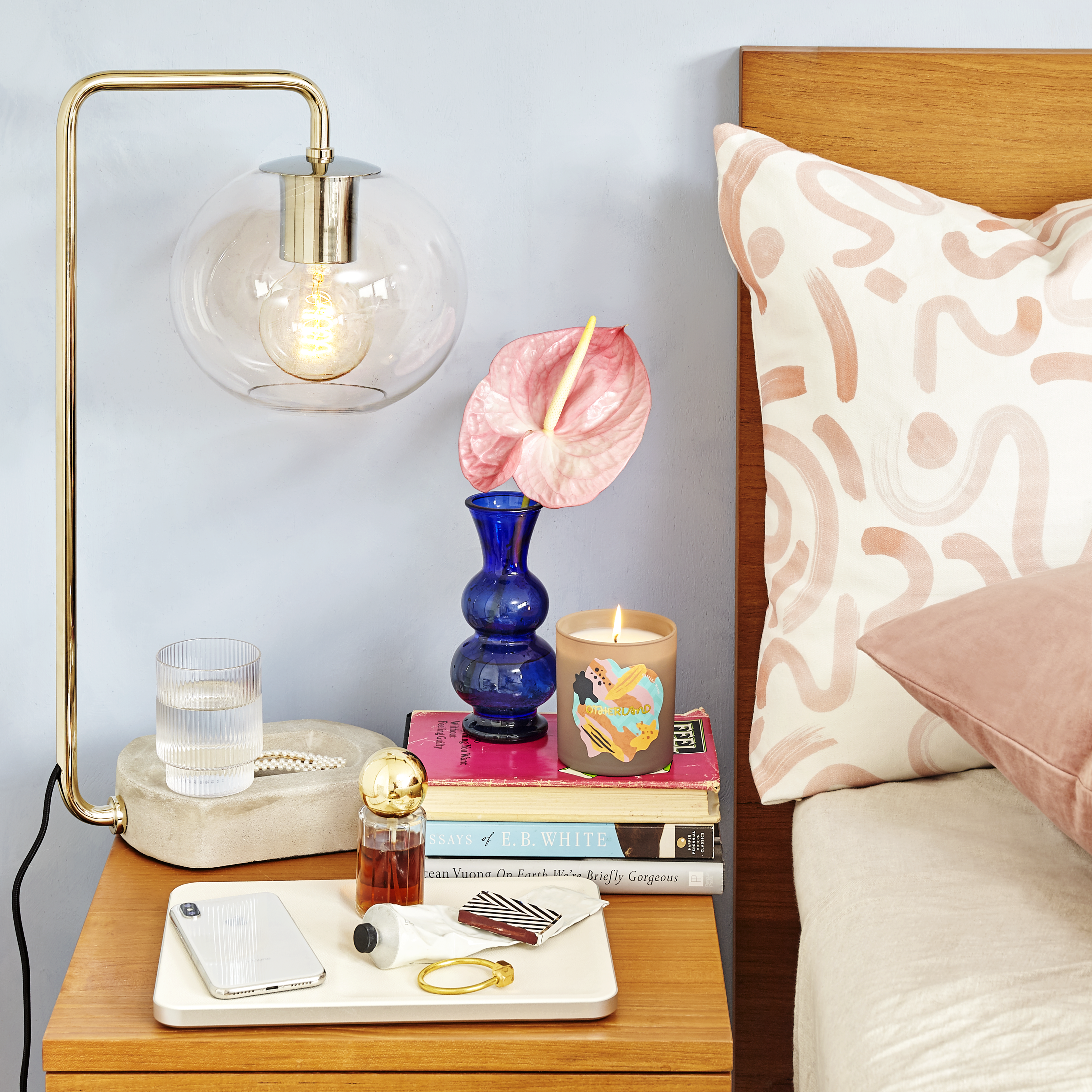Nightstand close-up with gold lamp and stacked books with pink tulip in blue vase by candle next to white bed with wooden backboard styled with a Pink Graphic Printed Pillow and a Pink Velvet Pillow