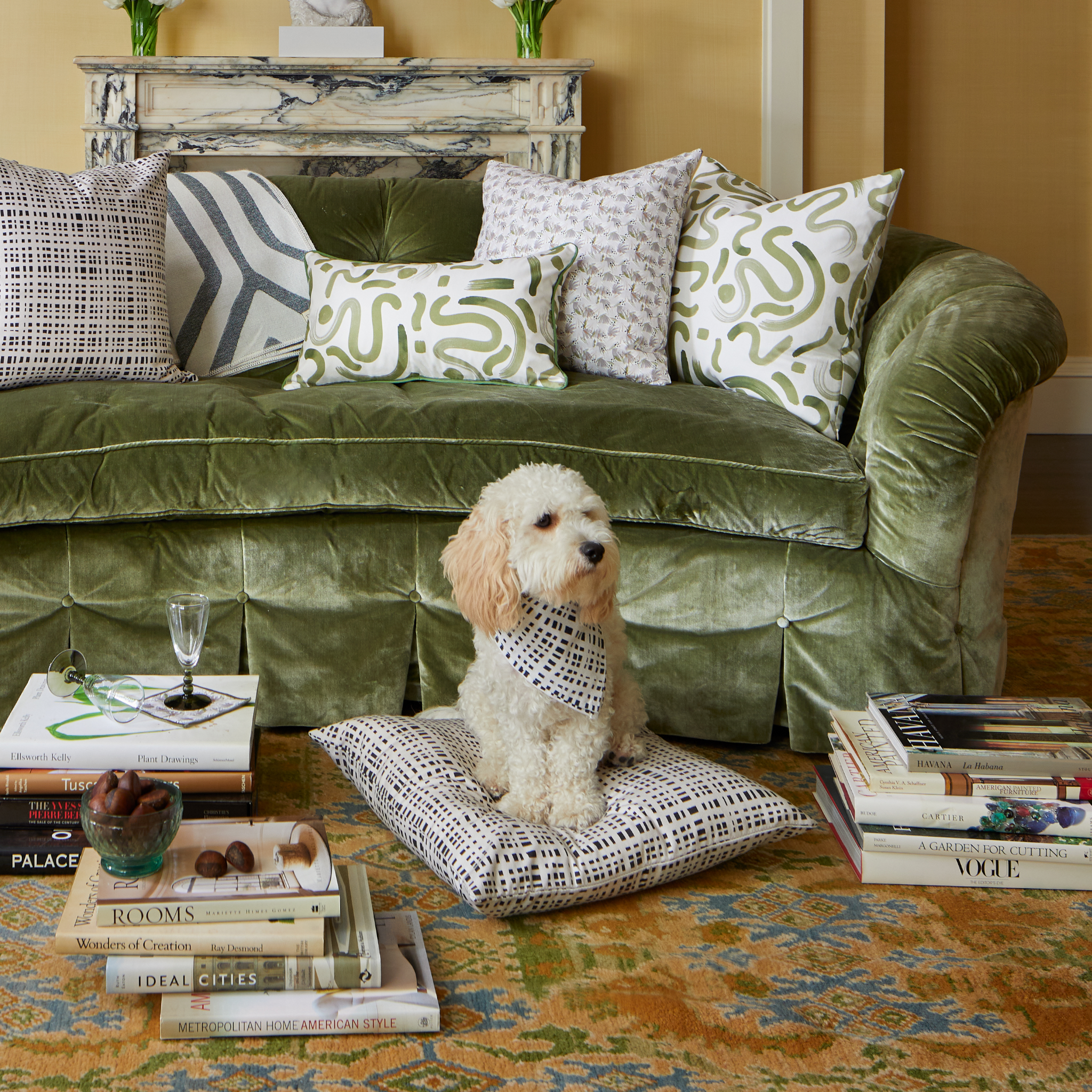 Living room styled with Black Gingham printed pillow, Grey Floral Printed Pillow, Moss Green Printed Lumbar and pillow on fern green velvet couch in the back of cream dog wearing Black Gingham fabric around neck on Black Gingham pillow surrounded by three stacks of books with decorations on top