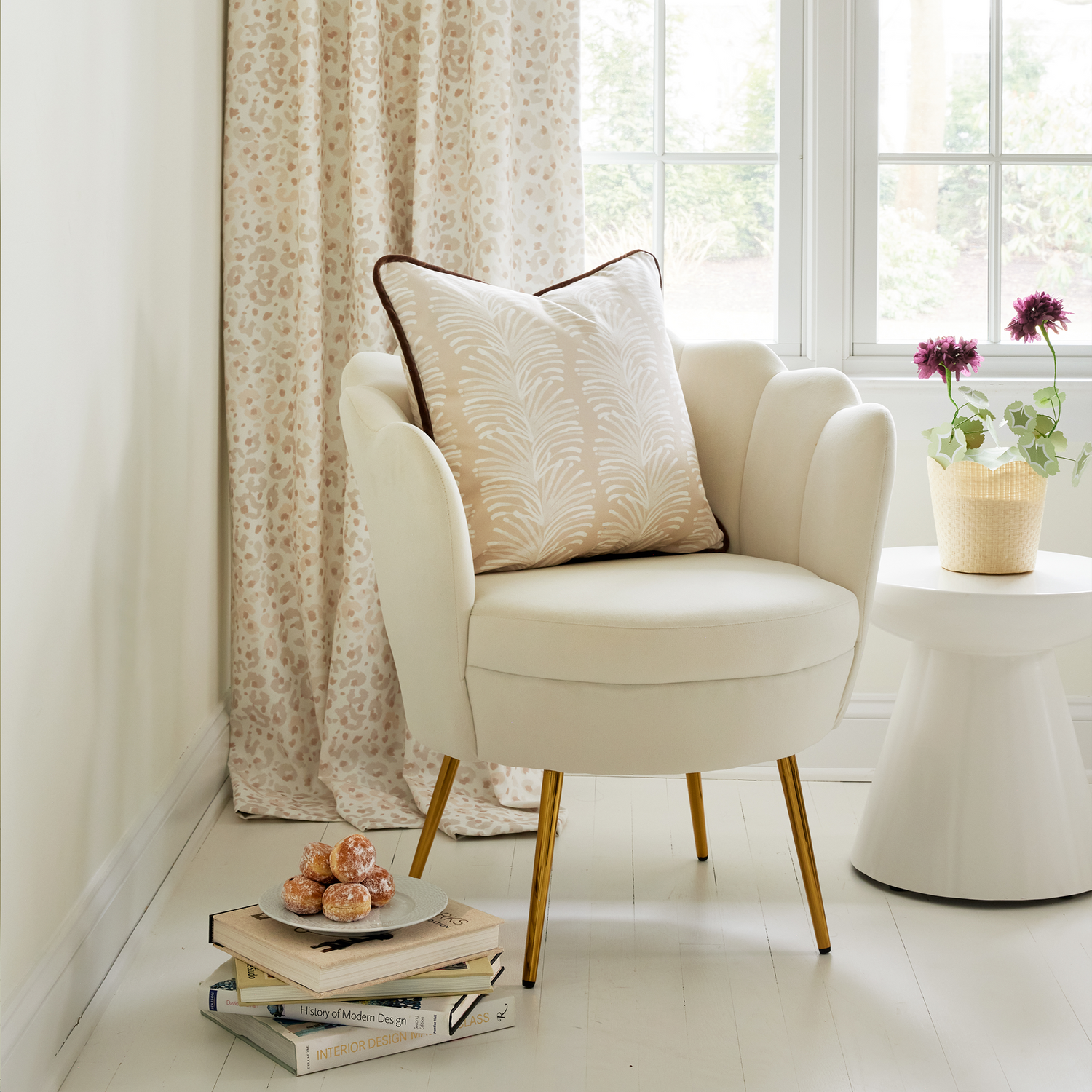 Window corner close-up styled with Beige Animal Print Curtains next to white sofa chair with a Beige Palm Printed Pillow by a small white circular table with flowers in clay pot on top. Stacked books on floor with dessert plate on top.
