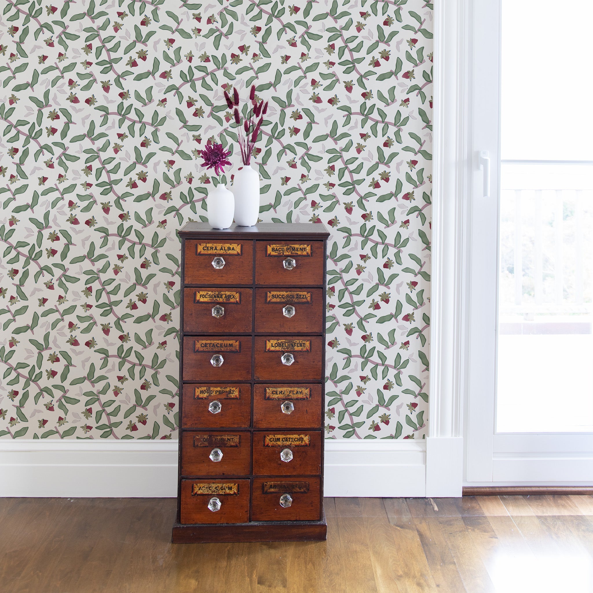 Strawberry & Botanical Printed Wallpaper with wooden cabinet in front and pink flowers on two white vases on top next to illuminated window