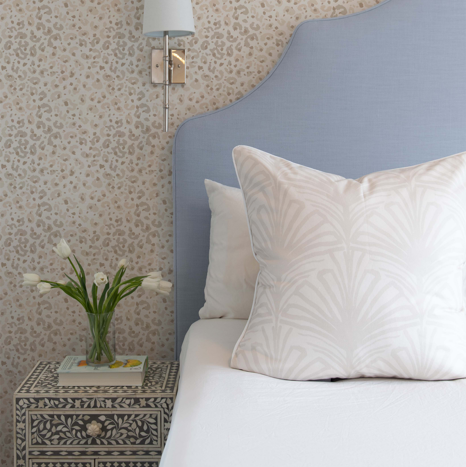Bed close-up styled with Beige Palm Printed pillow and Beige Animal Print Wallpaper by blue and white nightstand with white flowers in clear vase on top of book