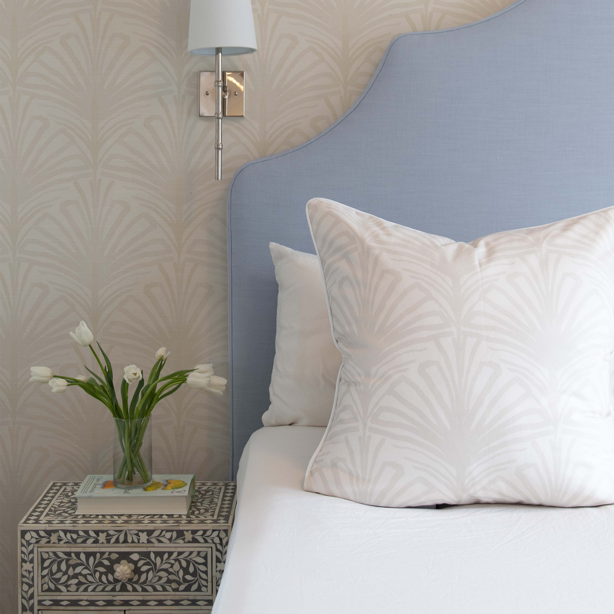 White bed close-up styled with Beige Palm Printed Pillow matching the beige palm printed wallpaper over white and blue nightstand with white tulips in clear vase on top of book