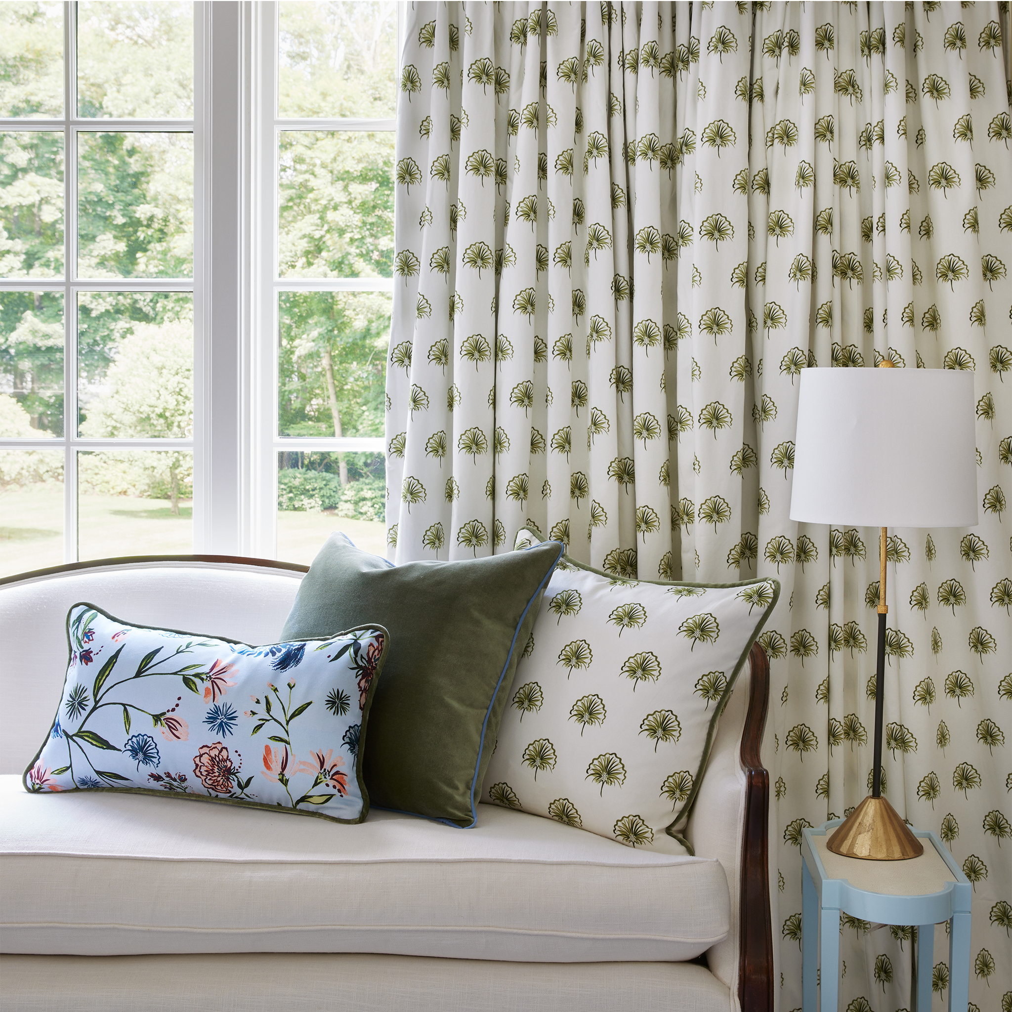 Living Room styled with Green Floral Printed Curtain and white couch in front with Blue Chinoiserie printed pillow, fern green pillow, and green floral printed pillow next to small baby blue table with gold and white lamp on top