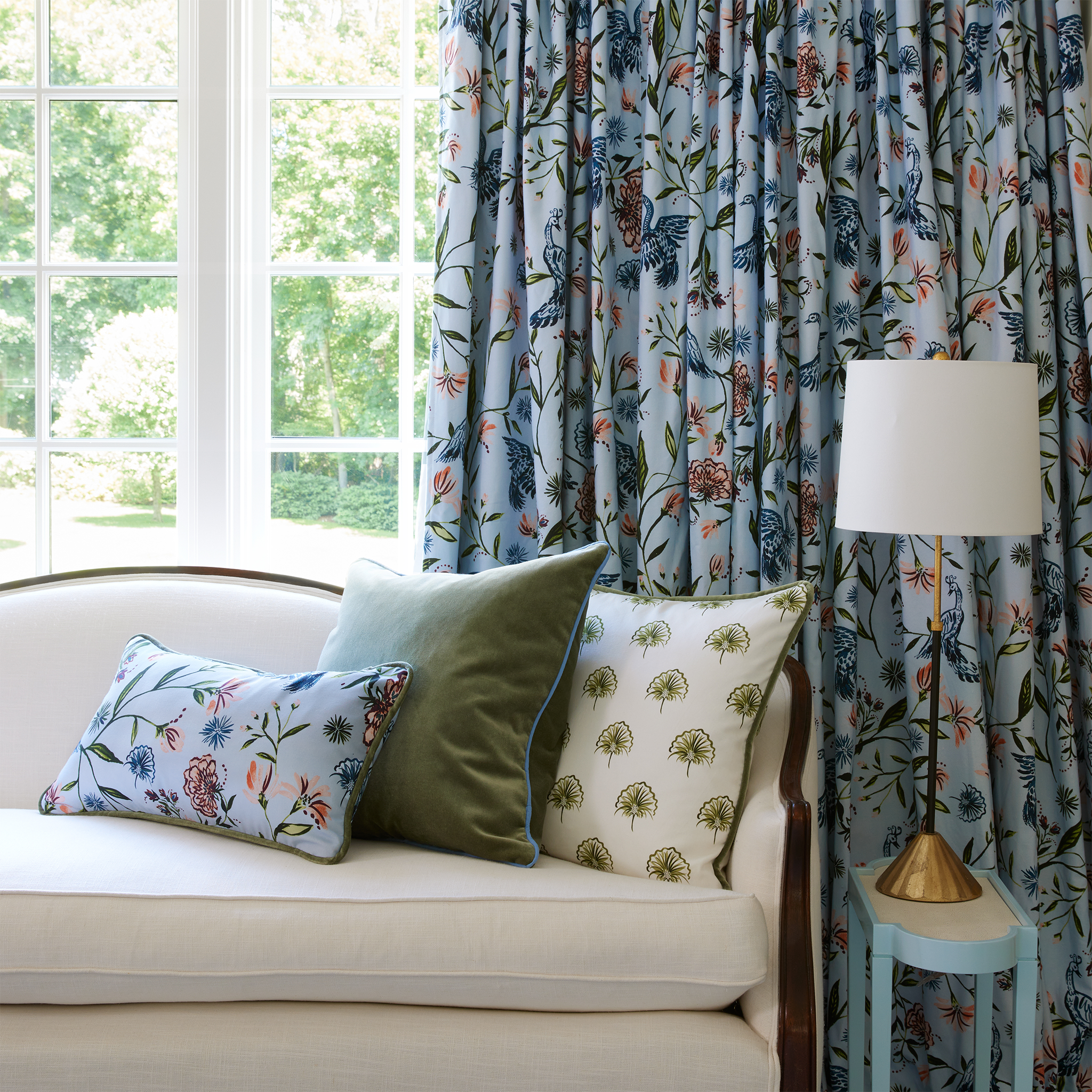 Living Room styled with Blue Chinoiserie Printed Curtain and white couch in front with Blue Chinoiserie printed pillow, fern green pillow, and green floral printed pillow next to small baby blue table with gold and white lamp on top
