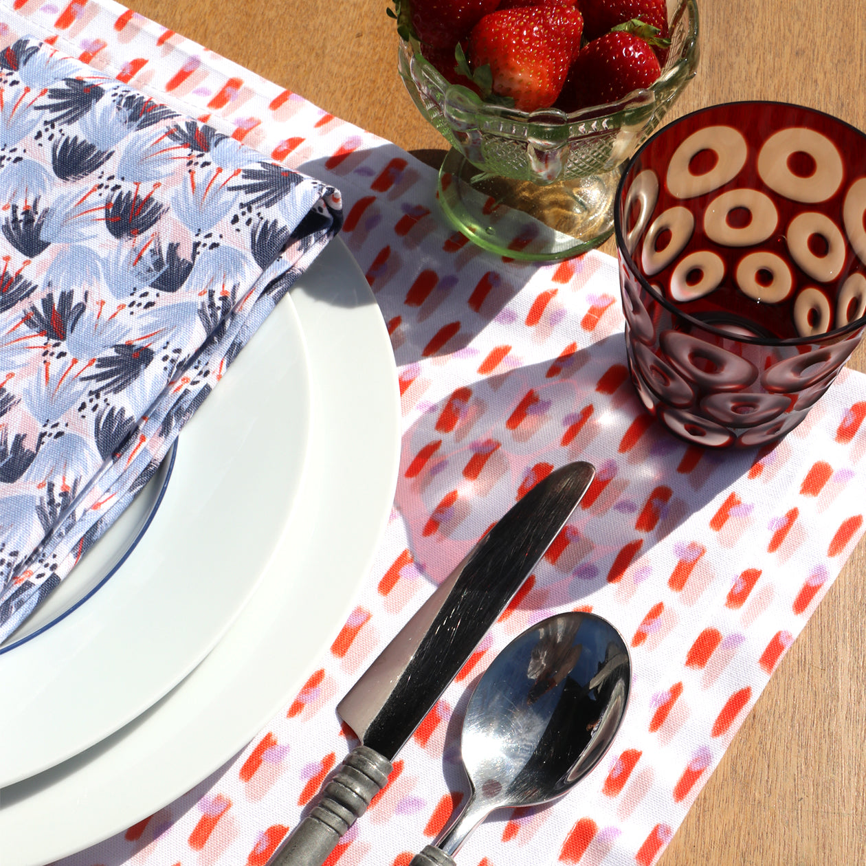 Light Pink Placemat close-up with red and blue printed napkin on top of two white plates by silverware and red glass by cup of strawberries