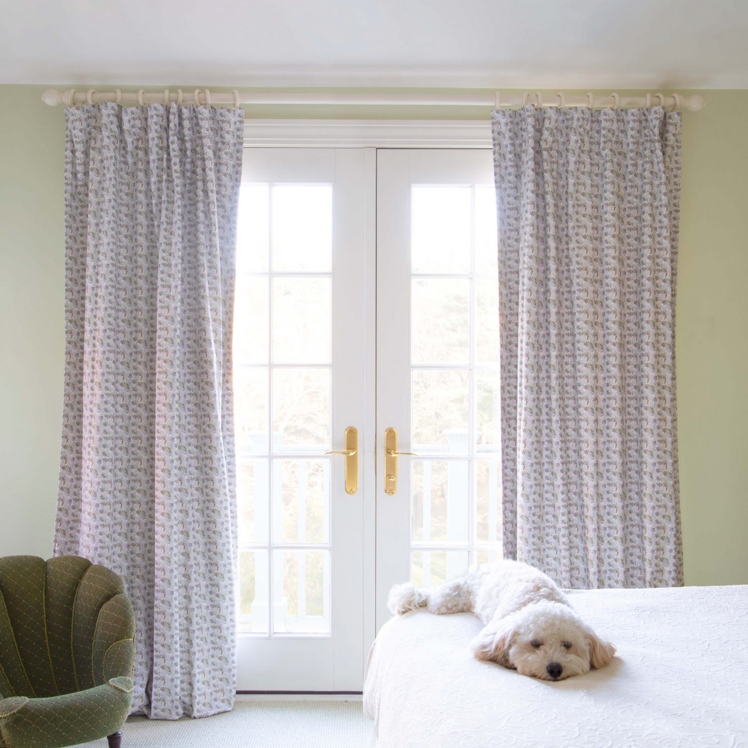 Bedroom styled with Grey Floral Printed Curtains on white rod in front on illuminated window by white dog on white bed next to fern green small sofa chair