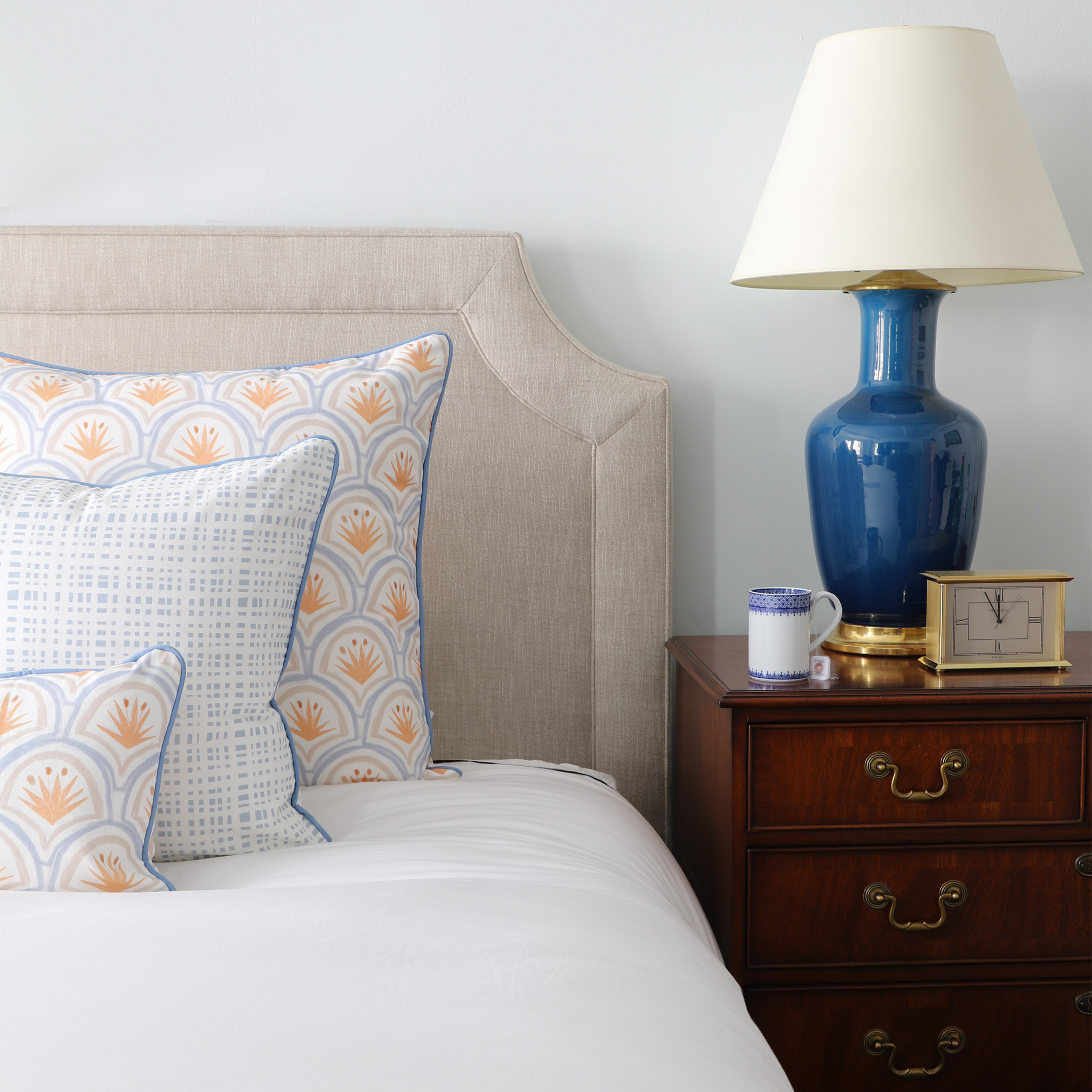 White bed close-up styled with Art Deco Palm Pattern Printed Pillow, Sky Blue Gingham Printed Pillow, and Sky Blue Gingham Printed Lumbar next to wooden nightstand with blue and white lamp on top by golden clock and tea cup