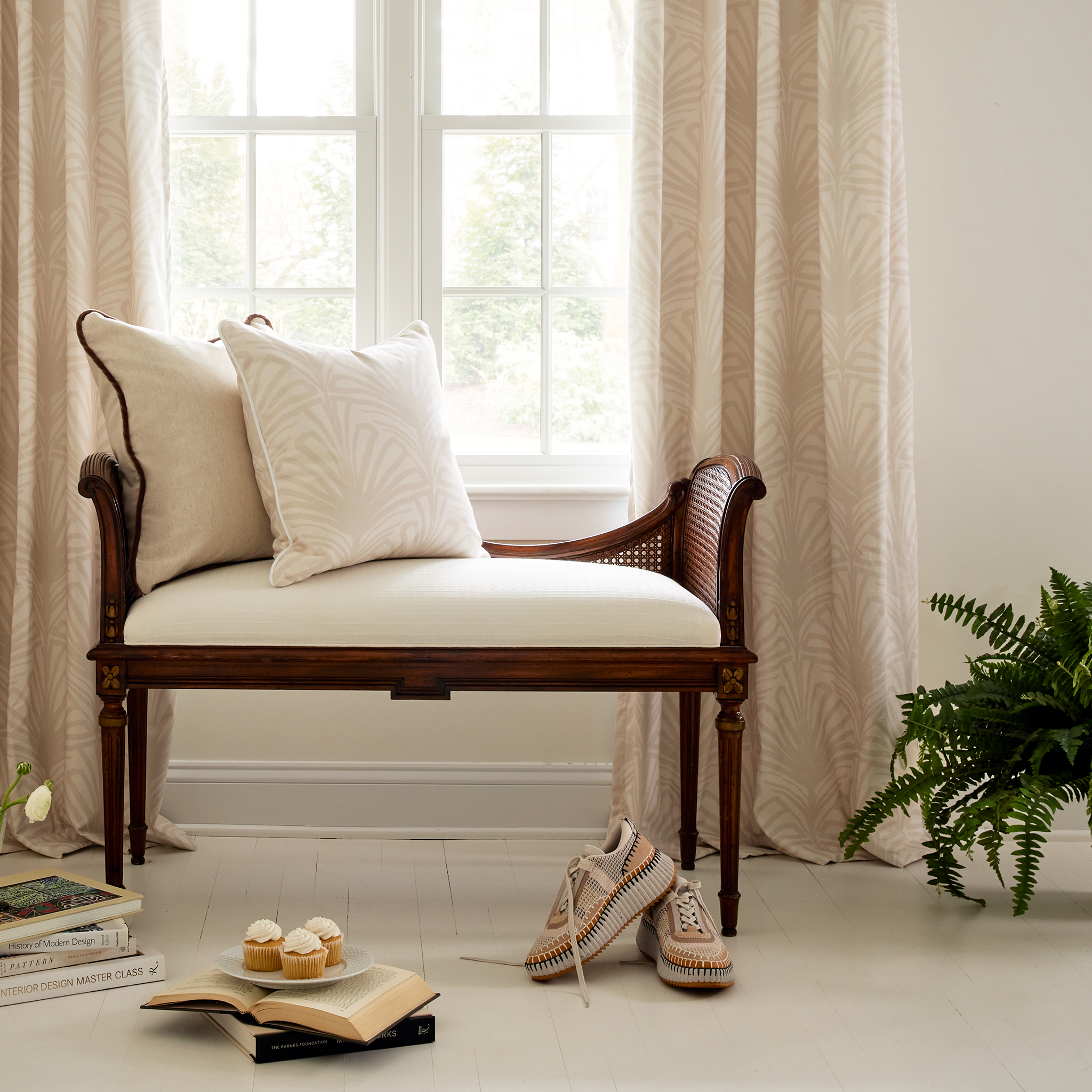 Close-up of window styled with Beige Palm Printed Curtains by wooden bench with Light Brown Linen Pillow and Beige Palm Printed Pillow next to plant on floor by tennis shoes and three cupcakes stacked on plate on two books