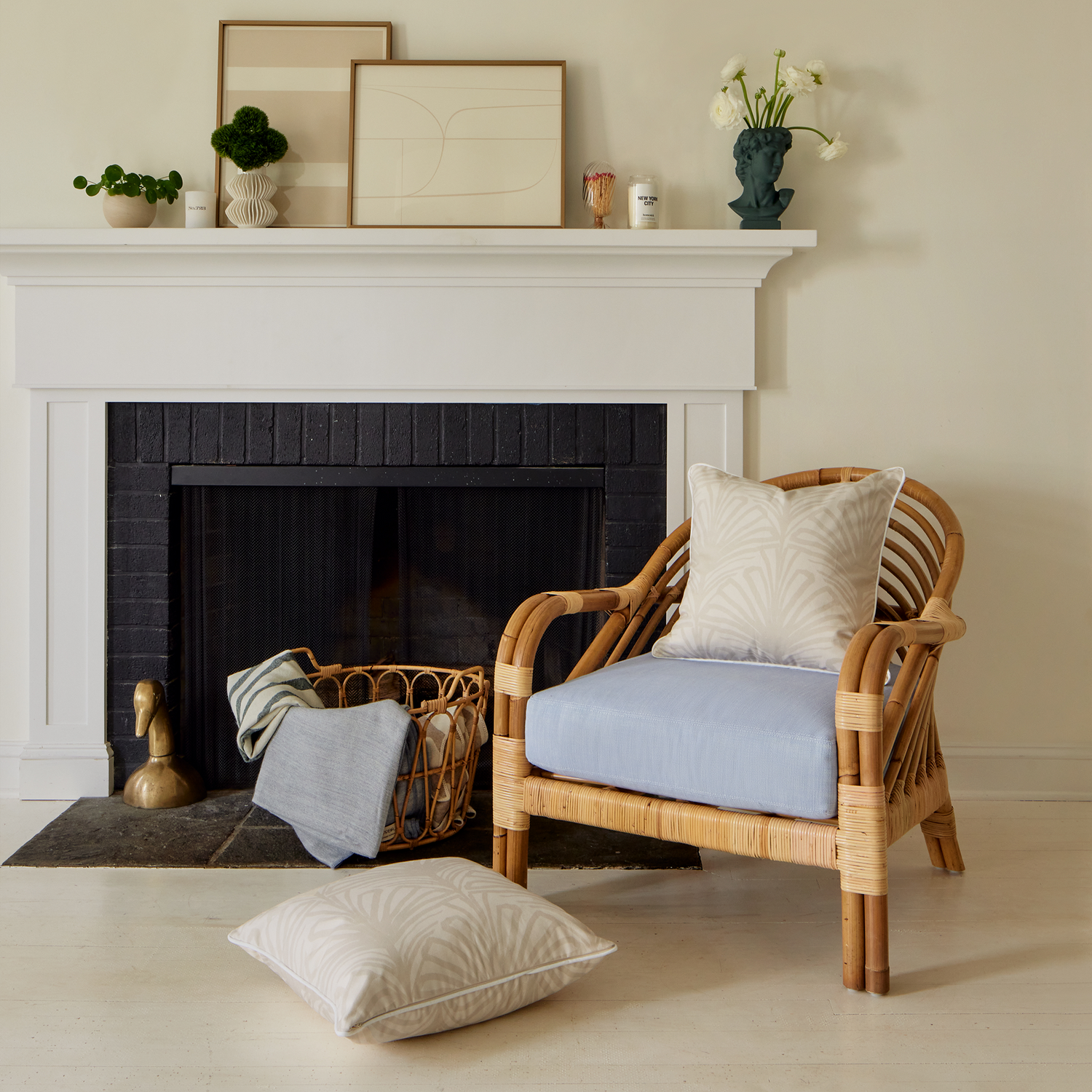 Fireplace styled with one beige palm printed pillow on wooden sofa chair and one beige palm printed pillow on the floor next to basket with blankets inside