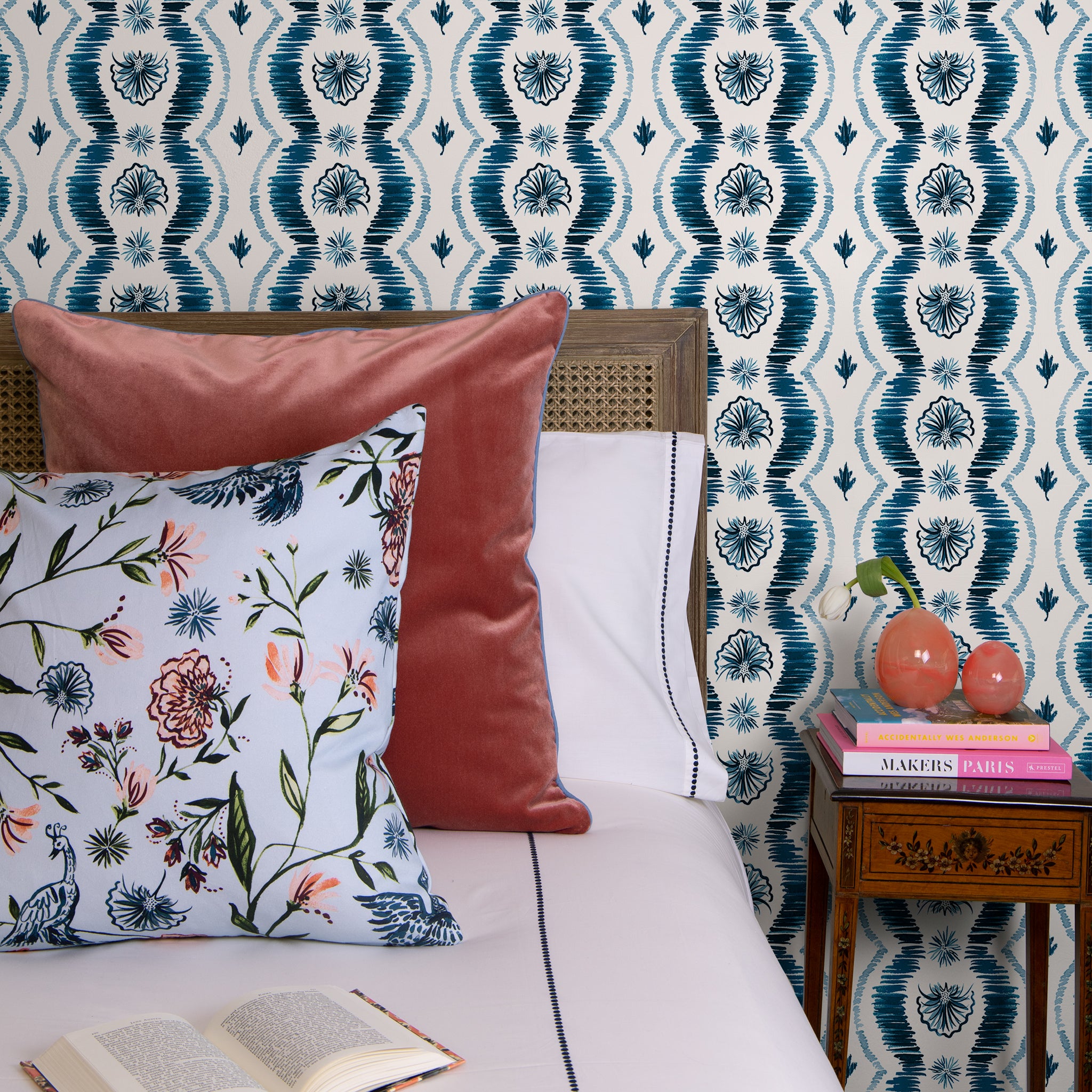 Blue Ikat Striped Pattern Wallpaper behind bed with one coral pillow, one daphne powder pillow, and a book next to a night stand with books and flower vase 