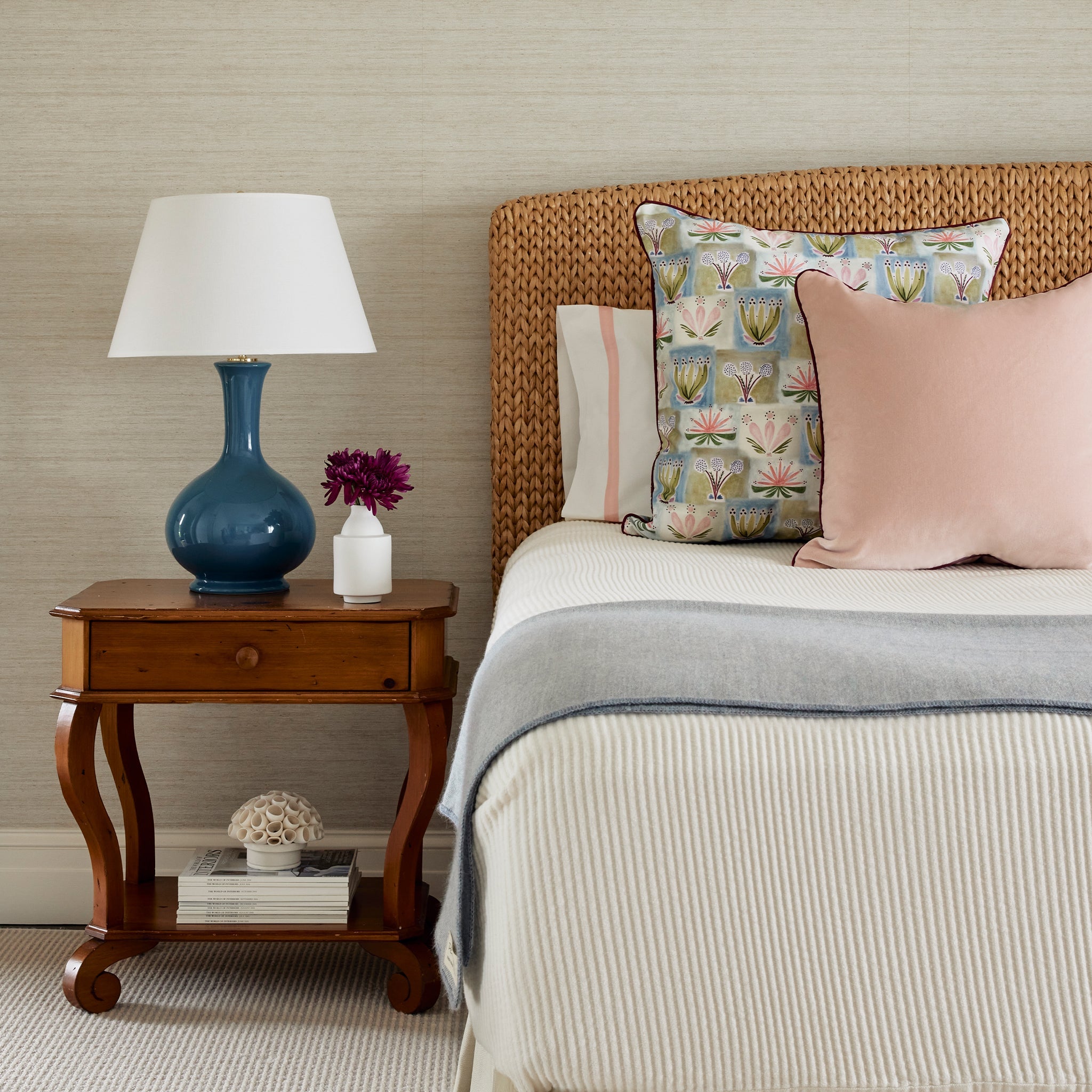 Bedroom corner styled with Hand-painted Floral Printed Pillow and Pink Velvet Pillow on white bed next to wooden nightstand with blue and white lamp on top by white vase with purple flowers