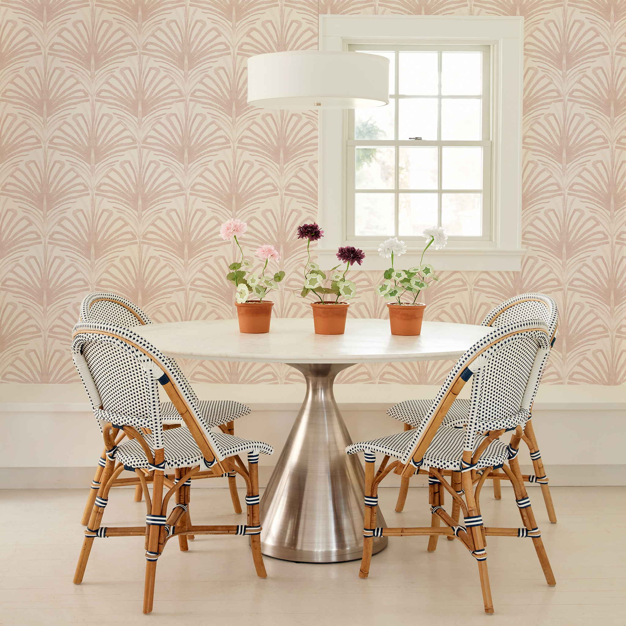 Circular table with flowers on three clay pots with 4 wooden chairs around it next to an illuminated window styled with a Rose Pink Palm Printed Wallpaper
