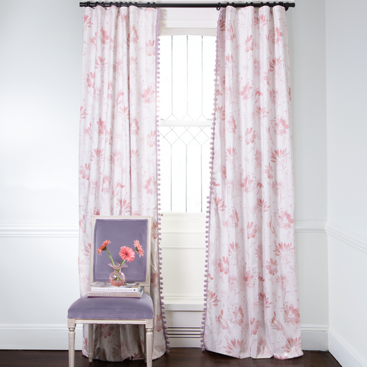 Pink Floral curtains with pink pom poms on metal rod in front of an illuminated window with lavender chair in front stacked with books and pink flowers on a vase