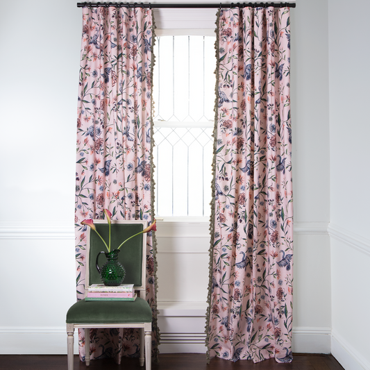 Pink Chinoiserie Printed curtains with Fern Green Pom Pom fringe on metal rod in front of an illuminated window with Green Moss chair with tulips in green vase on top of three stacked books
