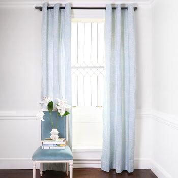 Powder Blue Abstract Printed Curtains on metal rod in front of an illuminated window with Sky Blue Velvet chair with white flowers in white vase on top of stacked books