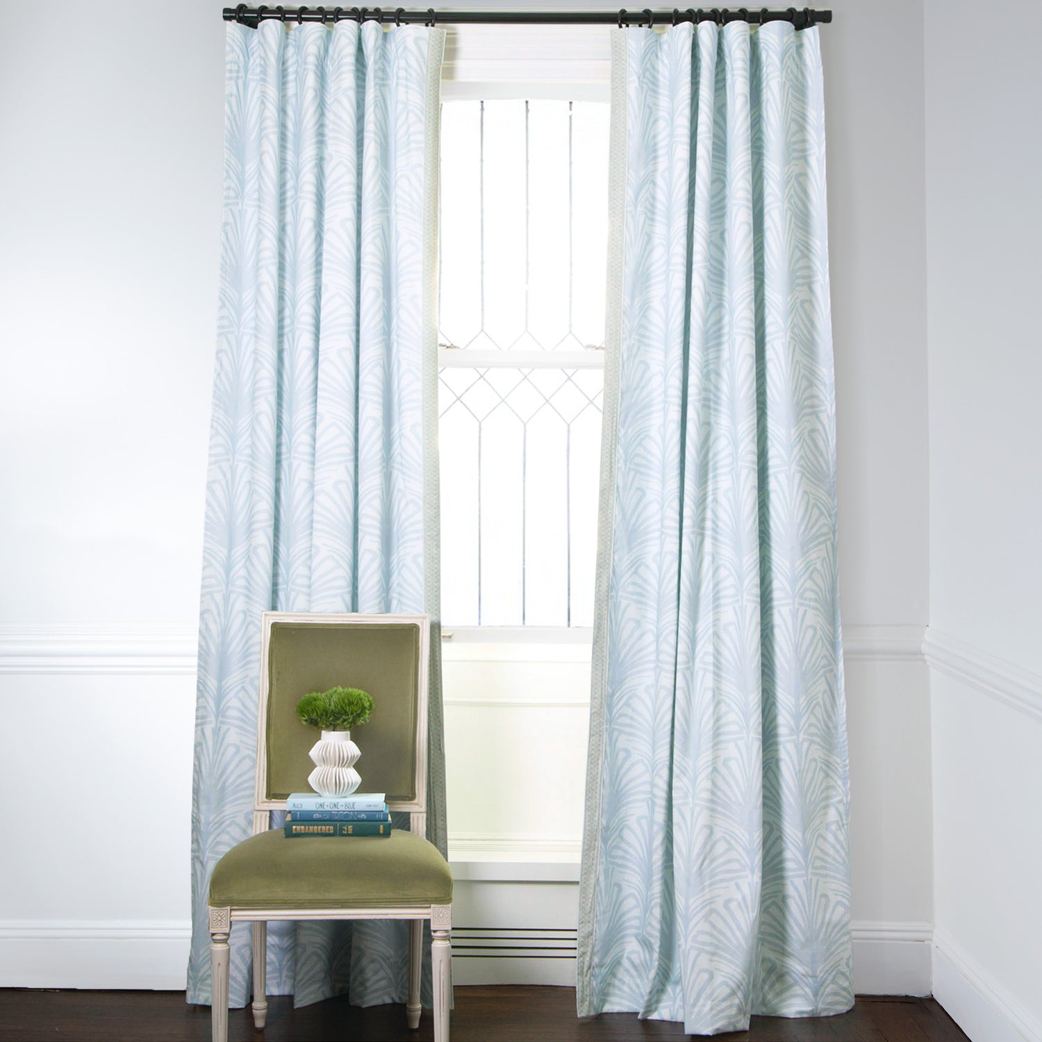 Sky Blue Palm Printed Curtains on metal rod in front of an illuminated window with Moss Green Velvet chair with plant in white vase on top of stacked books