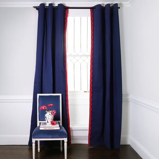 Navy Blue Curtains with red rick rack trim on metal rod in front of an illuminated window with Navy Blue Velvet chair with pink flowers in white and black vase on top of stacked books