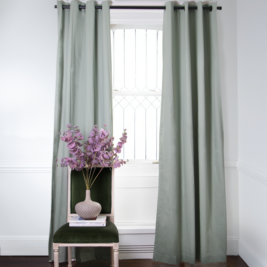 Sage Green Curtains on metal rod in front of an illuminated window with Fern Green Velvet chair with purple flowers in vase on top of stacked books