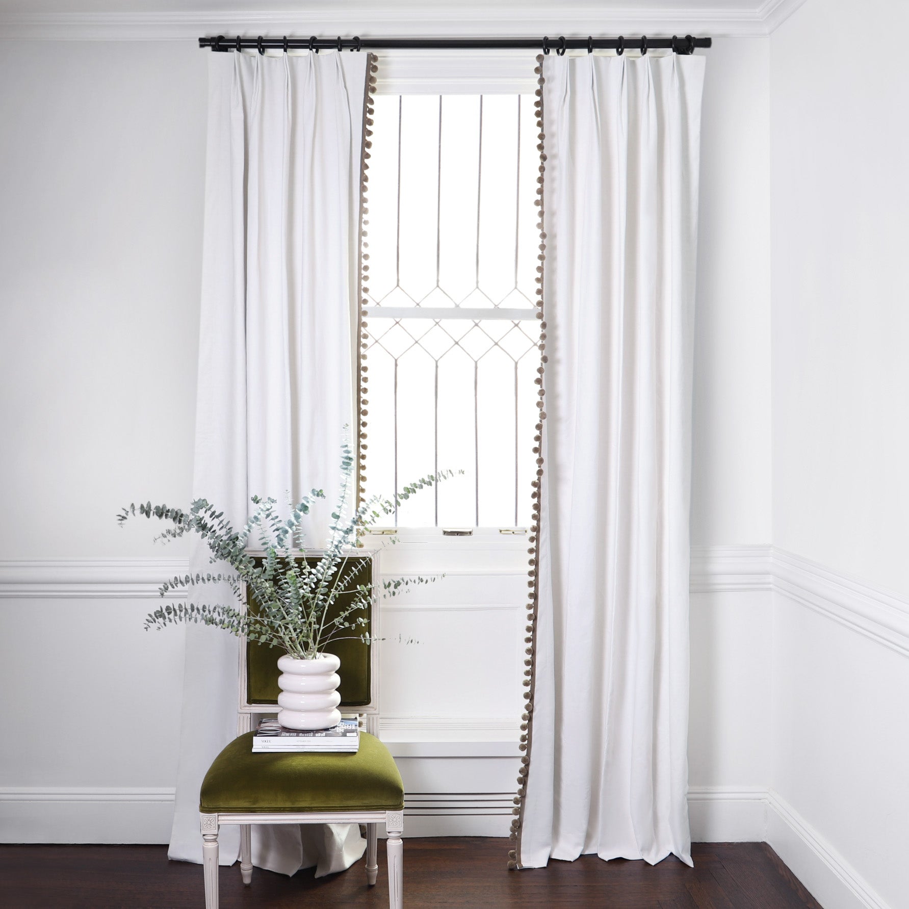 White Cotton Curtains with sage green pom poms on metal rod in front of an illuminated window with Sage Green Velvet chair with plants in white vase on top of magazines. 