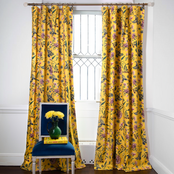 Daphne Canary printed curtains on metal rod in front of an illuminated window with navy chair in front stacked with a yellow book and yellow flowers on a green vase