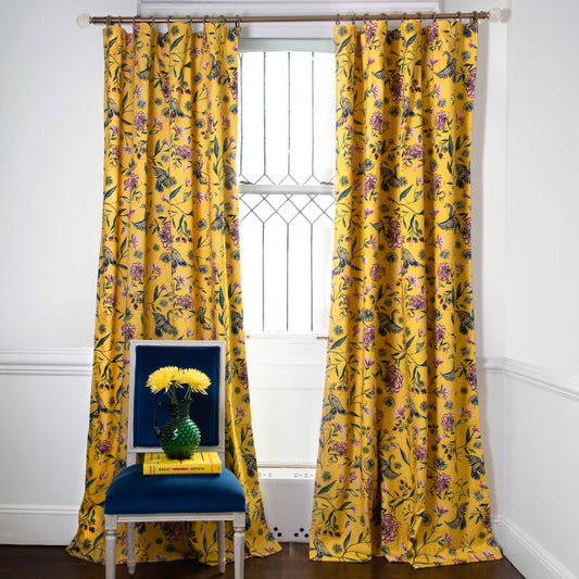 Daphne Canary printed curtains on metal rod in front of an illuminated window with navy chair in front stacked with a yellow book and yellow flowers on a green vase