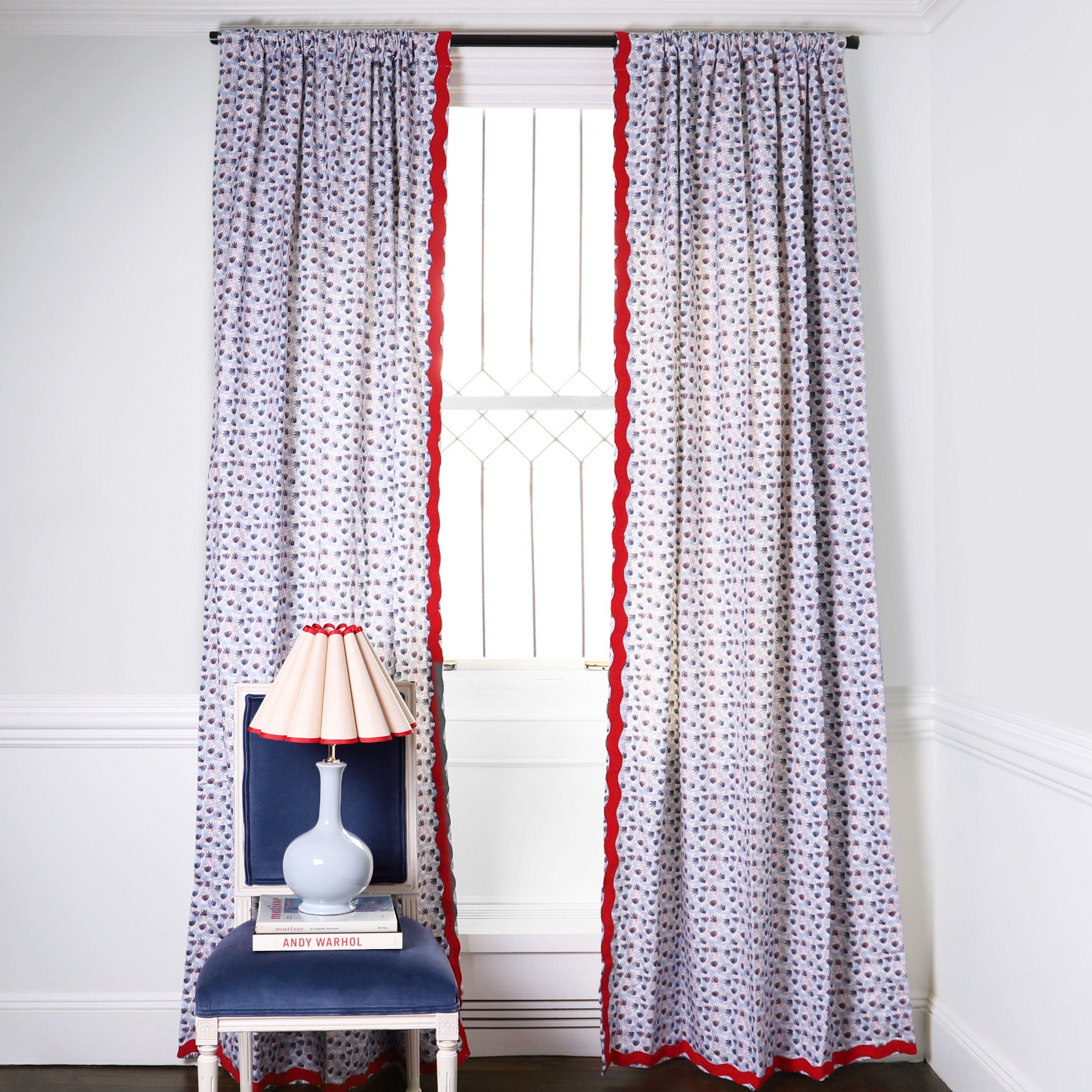 Red and Blue Printed Curtains with red fringe on metal rod in front of an illuminated window with Navy Blue Velvet chair with baby blue, cream and red lamp on top of a book
