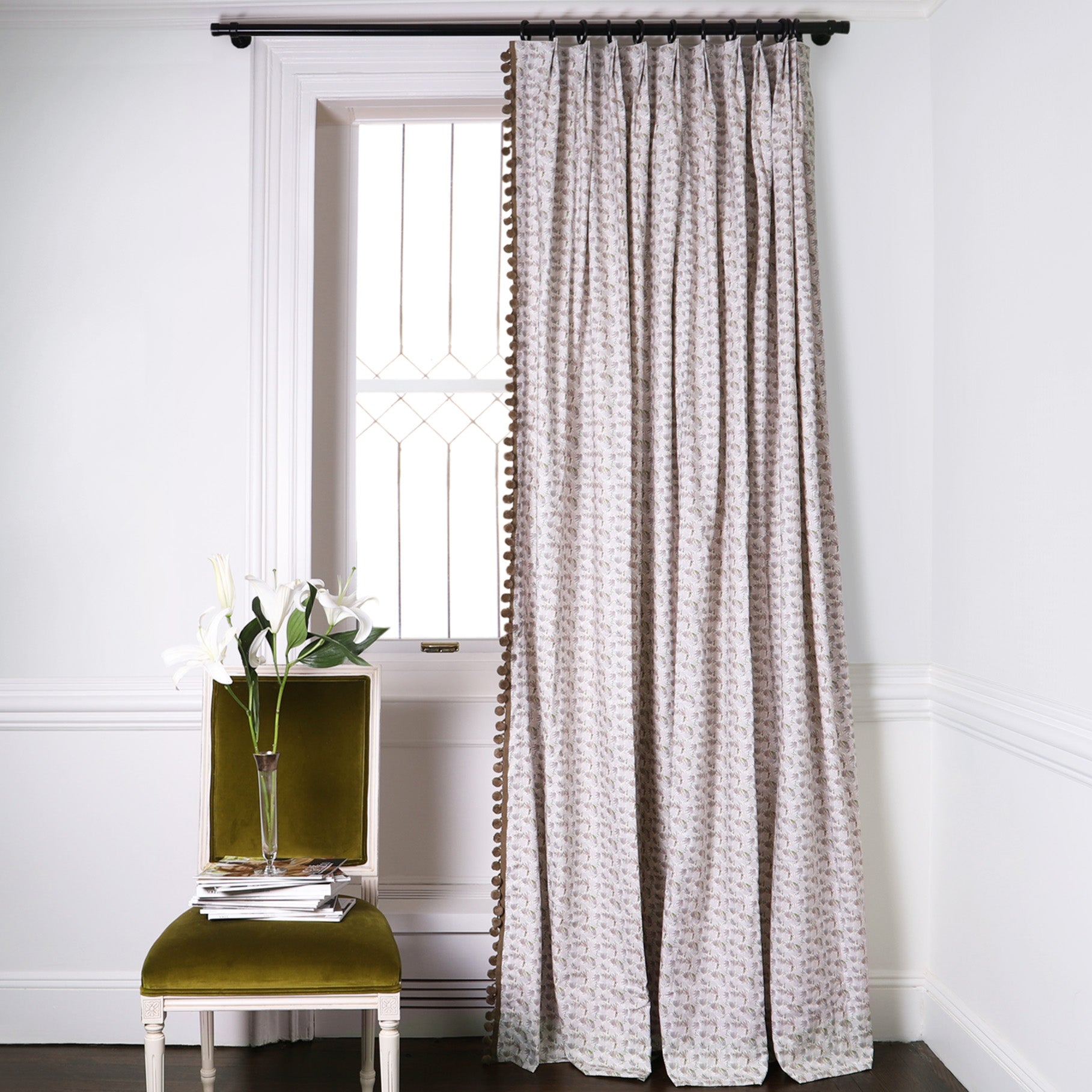 Grey Floral Printed Curtains with brown pom pom fringe on metal rod in front of an illuminated window with Green Moss Velvet chair with tulips on vase on top of stacked magazines